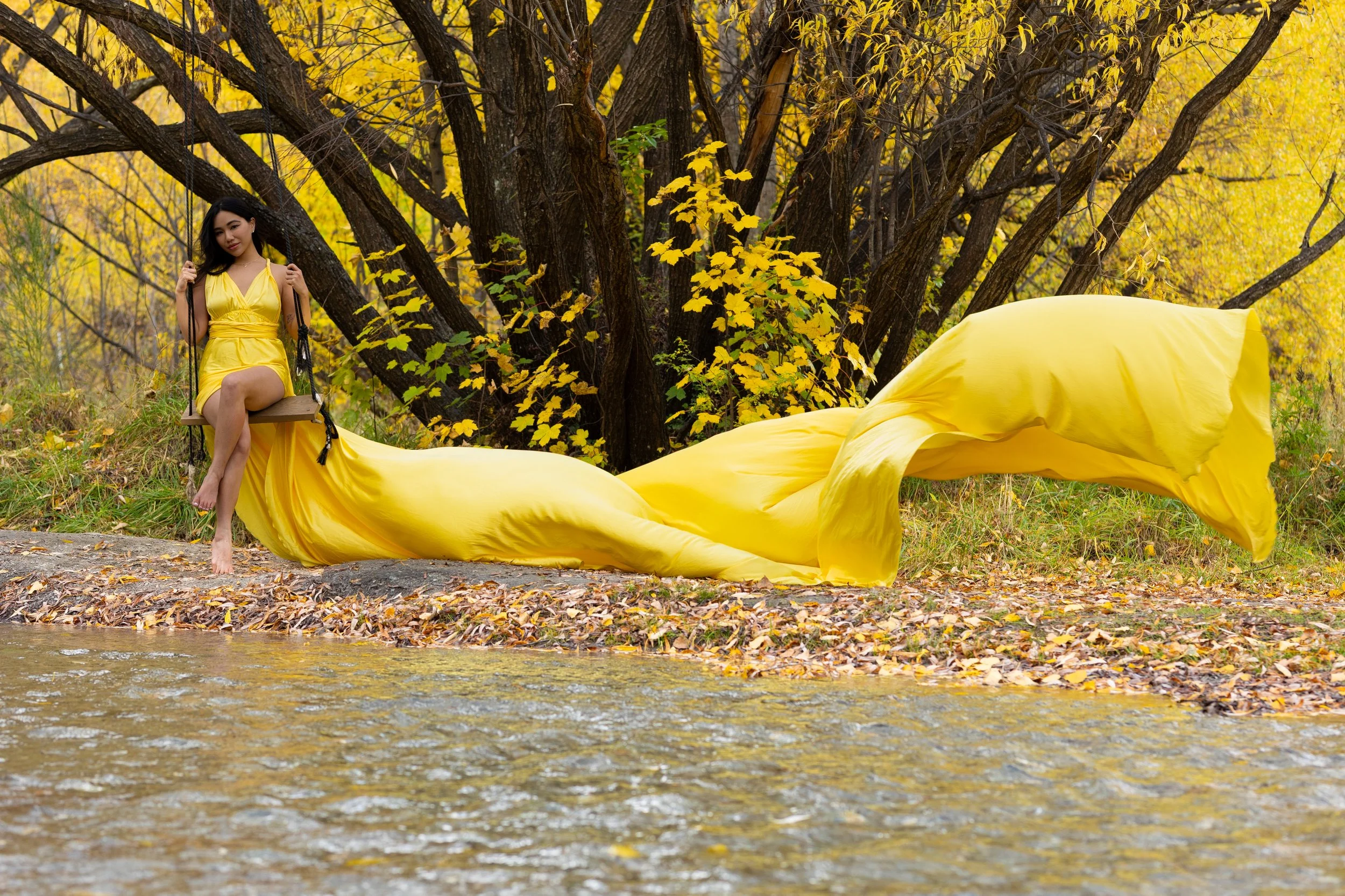 A woman in a yellow dress sitting on a swing in a forest with autumn foliage, a yellow fabric flows on the ground, and a yellow umbrella is nearby.