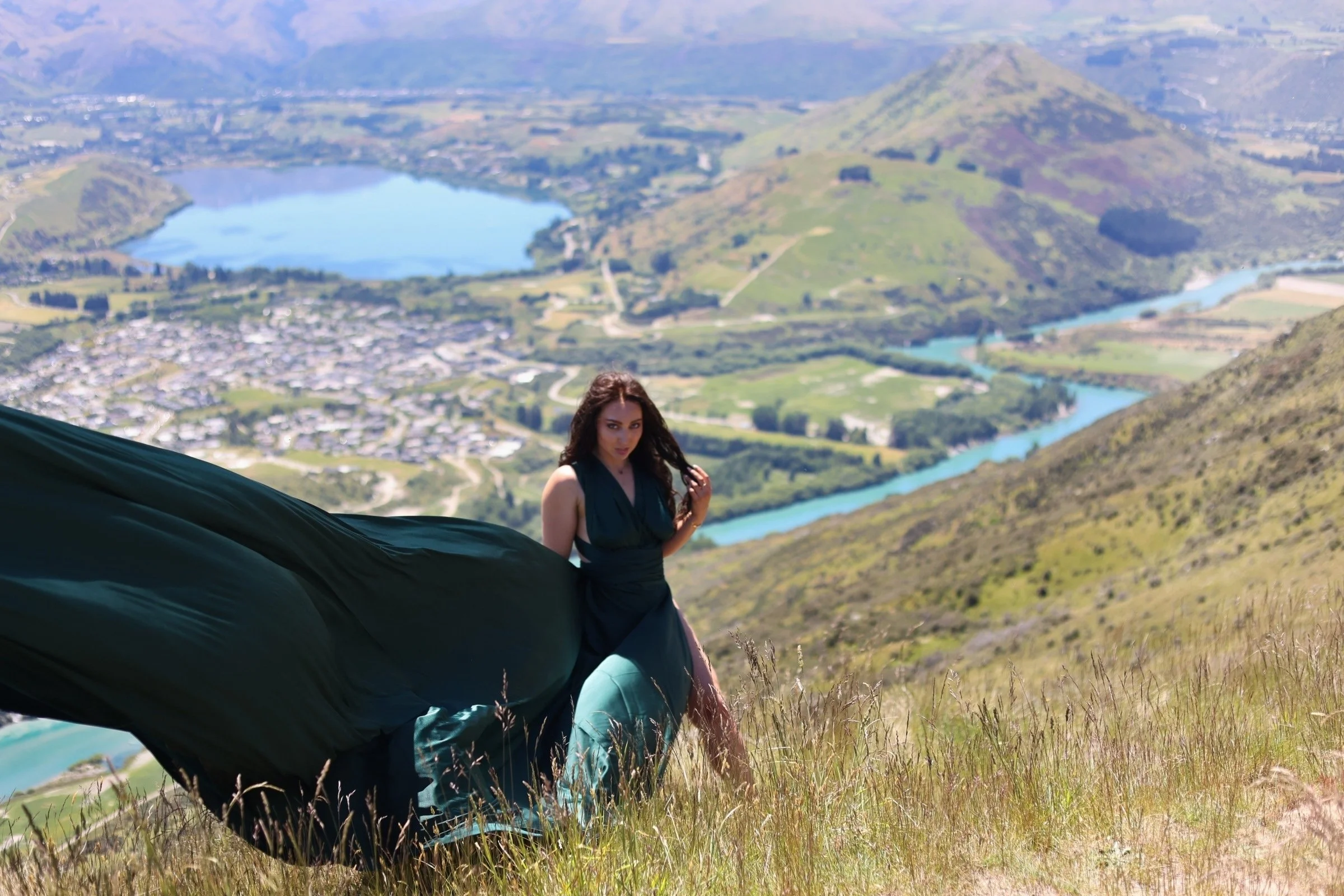 A woman in a dark dress standing on a grassy hillside with a scenic view of lakes, mountains, and a small town in the background.