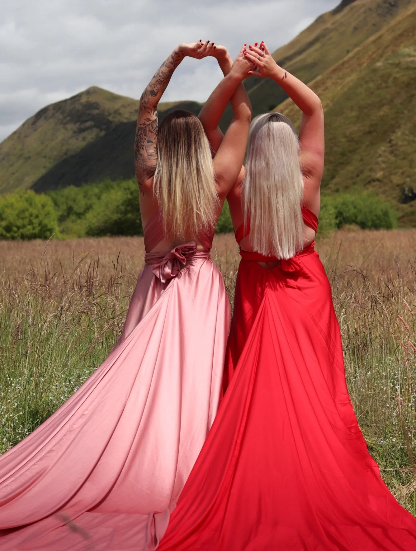Two women in long, flowing dresses, one pink and one red, standing in a field with mountains in the background, holding their arms above their heads.