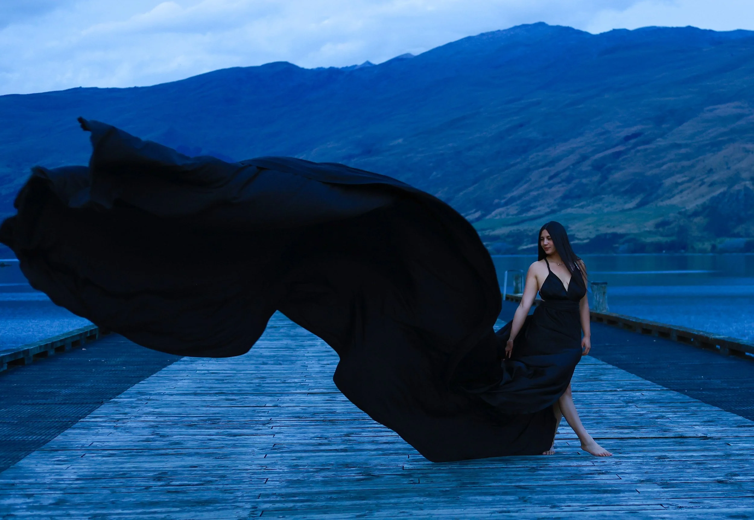A woman in a long black dress kneels on a wooden dock near a body of water, with mountains in the background during twilight. A large black cloth or fabric is being waved or blown in the foreground.