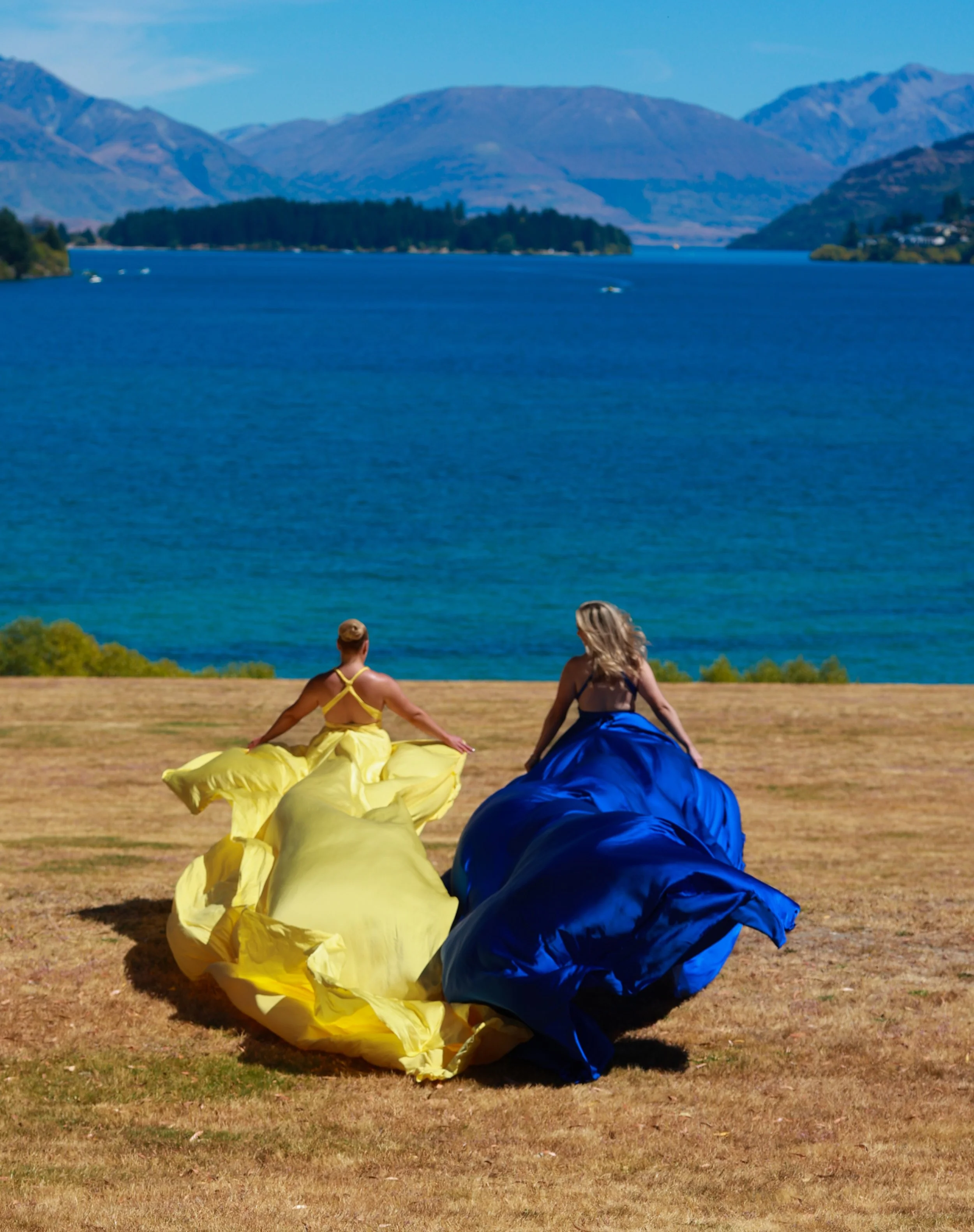 Two women sitting on a grassy field near a lake with mountain ranges in the background. They are dressed in colorful dresses, yellow and blue, and appear to be enjoying a sunny day.