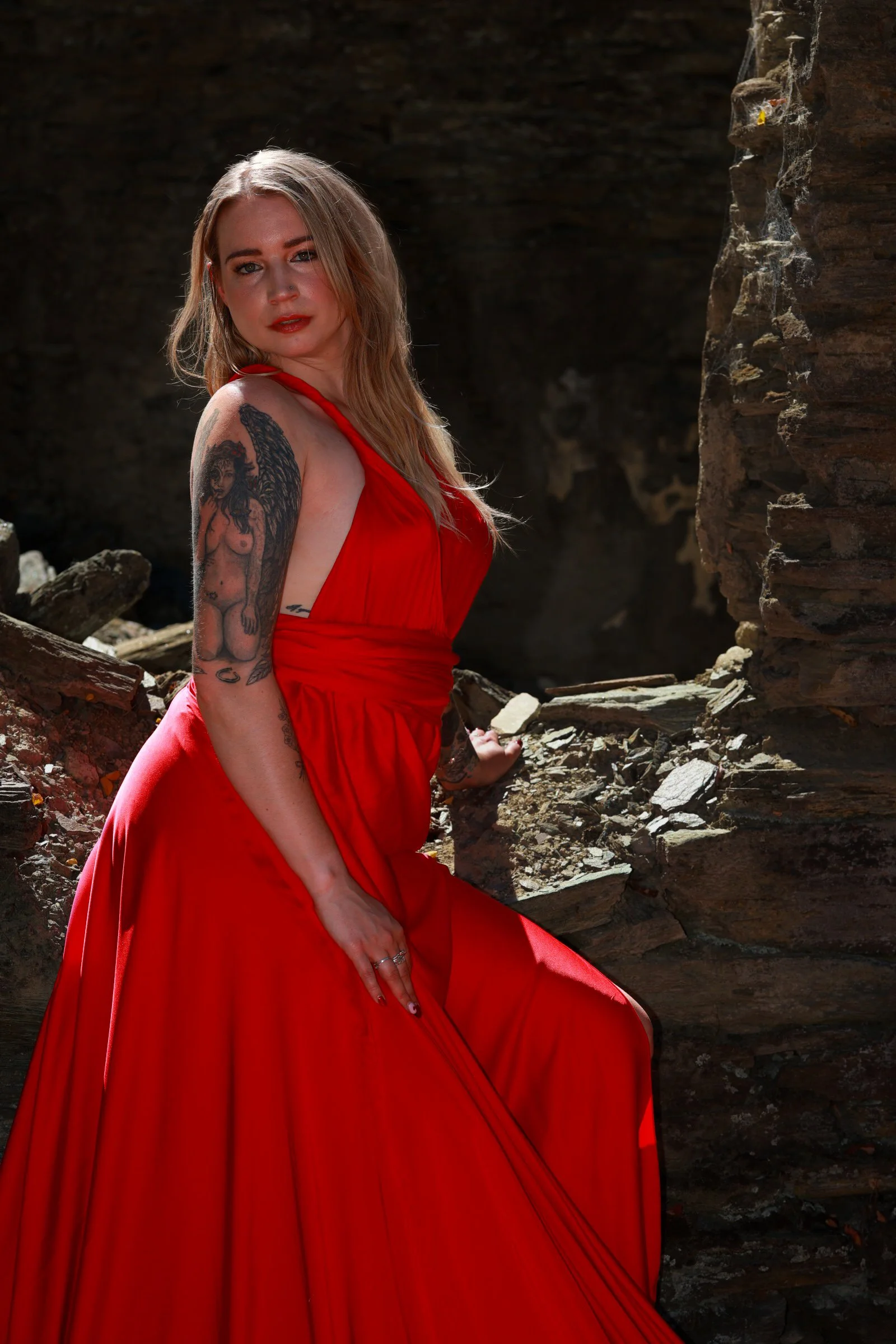 A woman in a red dress sitting near rocky terrain and a cliff with a tattoo on her arm.