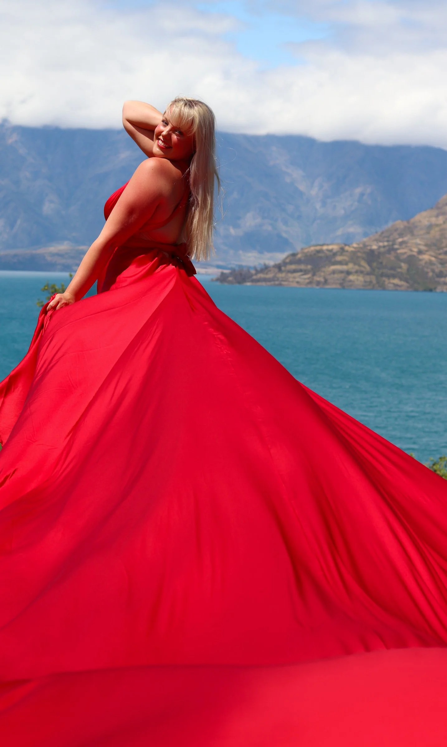 A woman in a red dress smiling and posing outdoors with a mountainous lake background.
