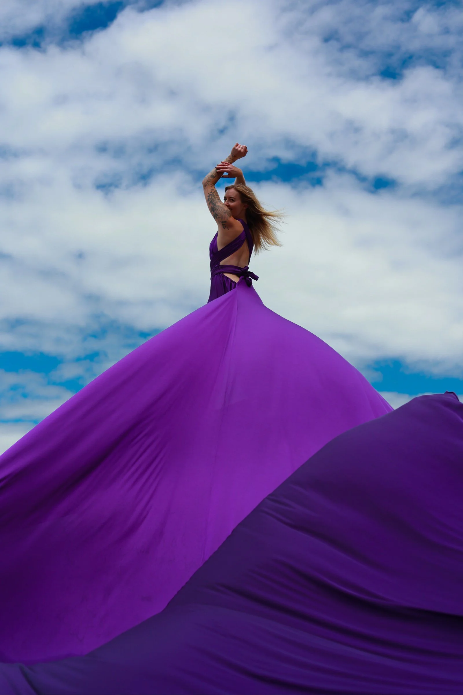 A woman in a purple dress with tattoos on her arm, standing on a purple fabric structure against a partly cloudy sky.