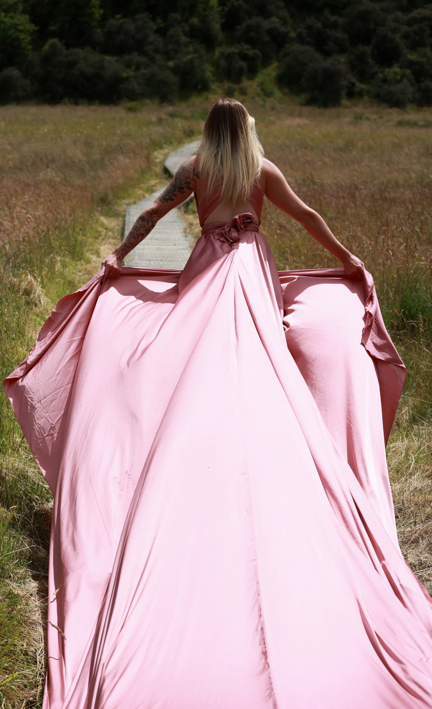 Woman in a flowing pink gown standing on a wooden path in a grassy field, with trees in the background, seen from behind.