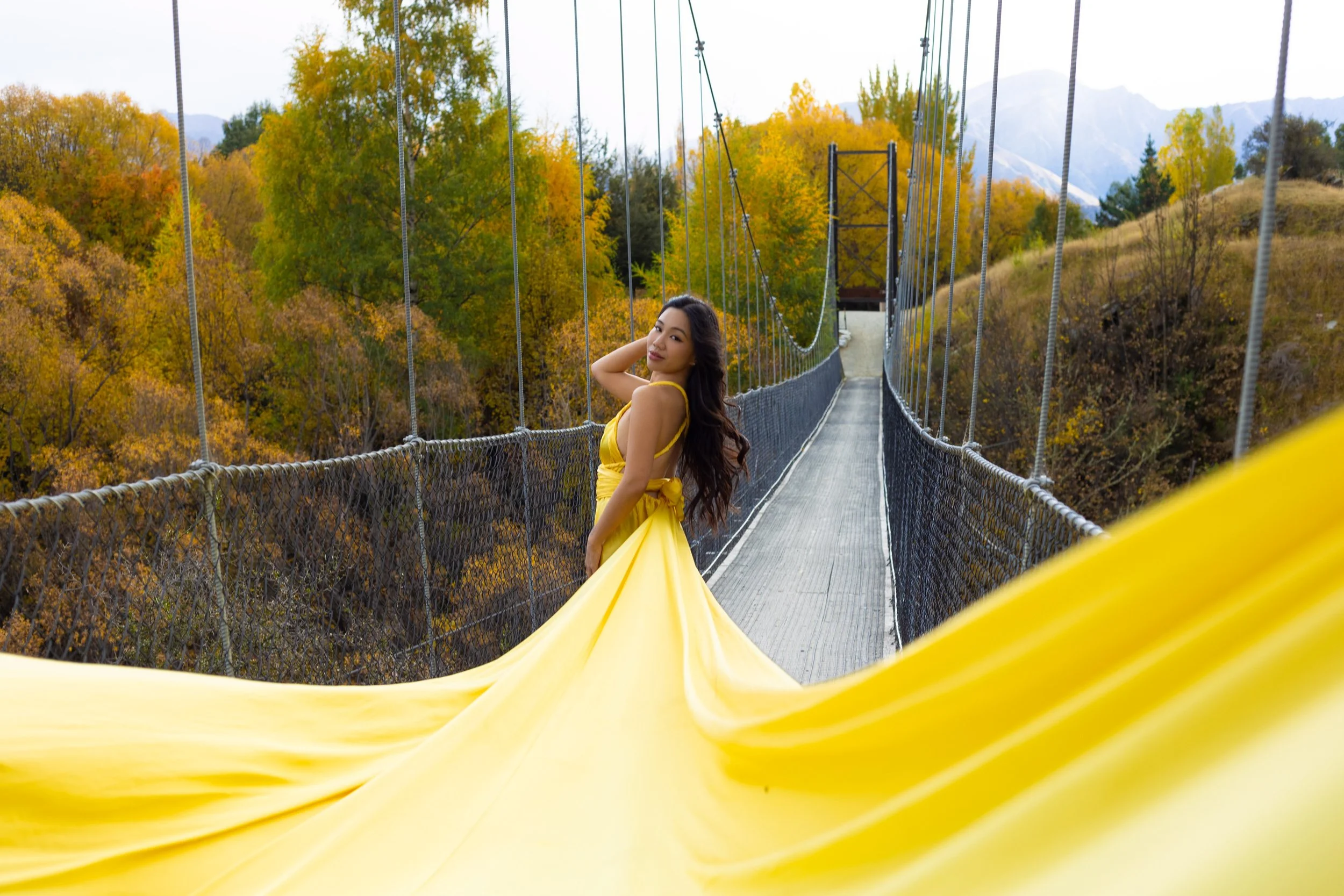 A woman in a yellow dress sitting on a yellow slide on a suspension bridge, surrounded by trees with autumn foliage and mountains in the background.