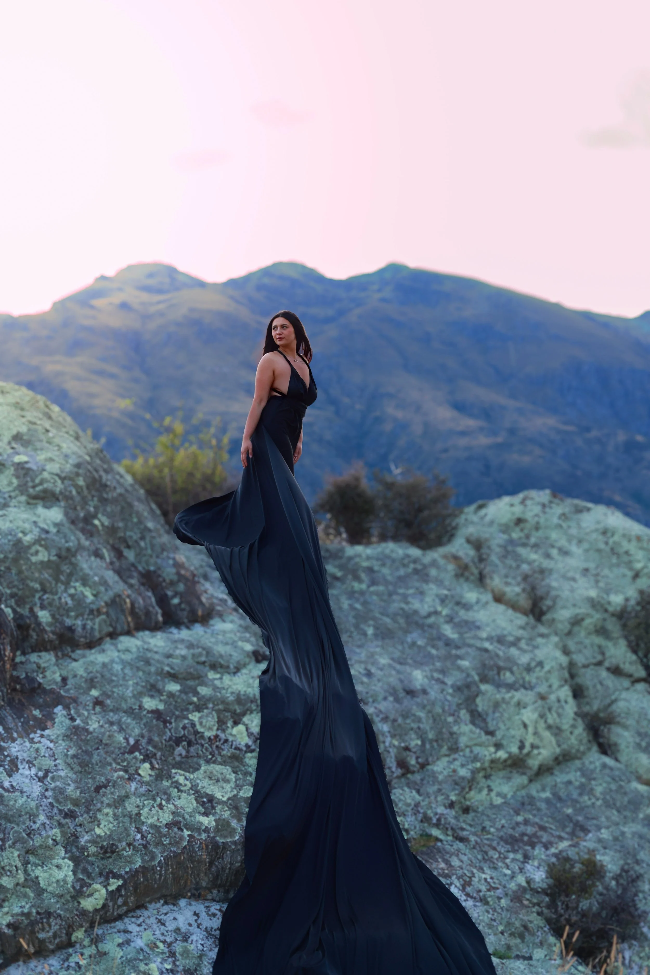 A woman in a long, flowing black dress standing on large moss-covered rocks outdoors with mountains in the background at sunset or sunrise.