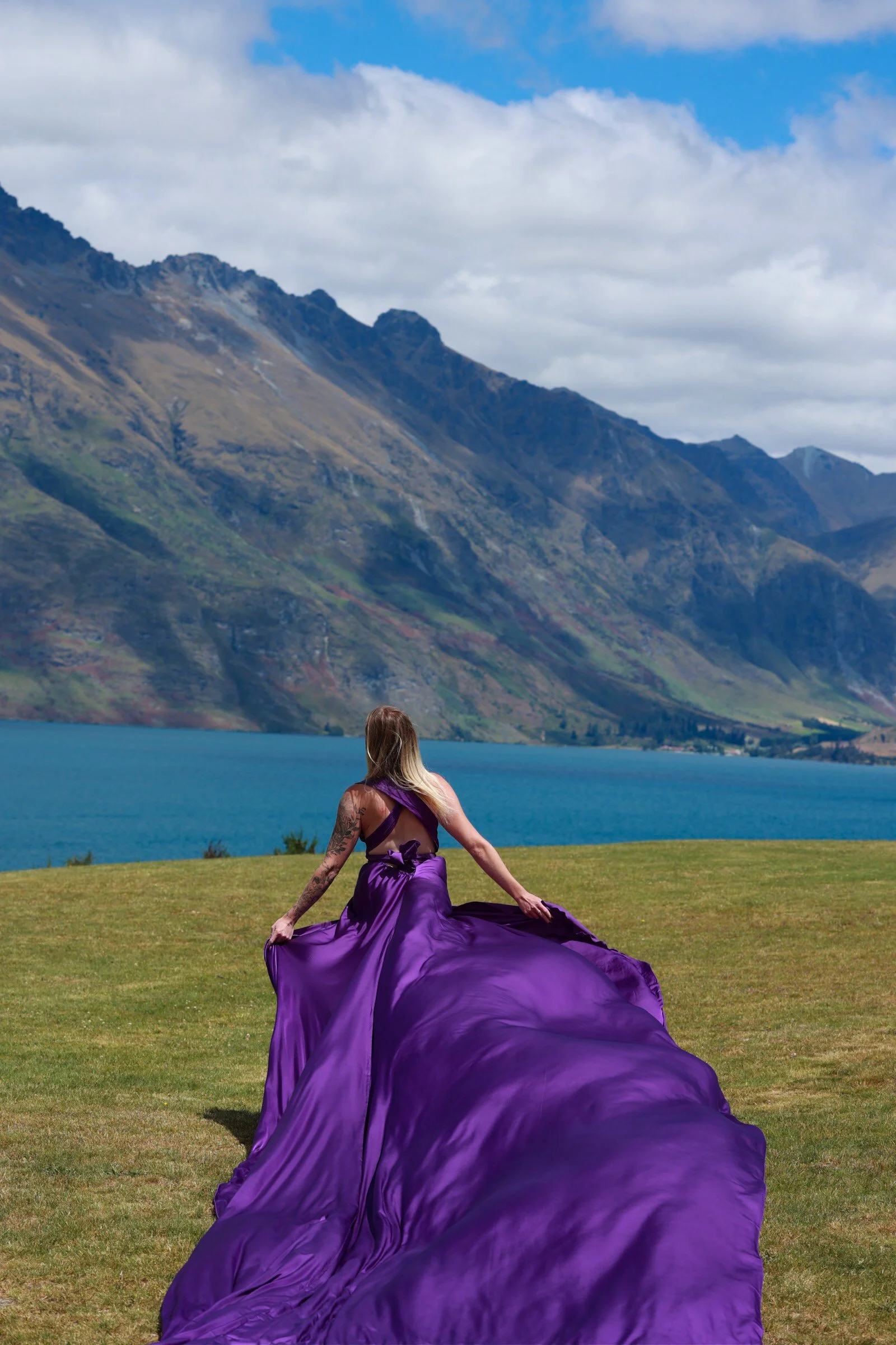 Woman in a flowing purple dress sitting on a grassy field near a lake with large mountains in the background and cloudy sky.