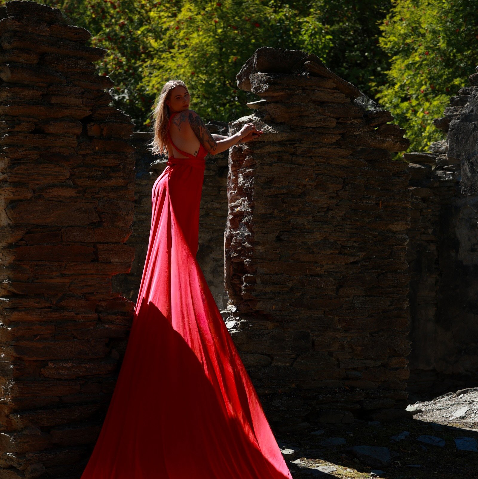 A woman in a red gown standing among stone ruins with green foliage in the background.