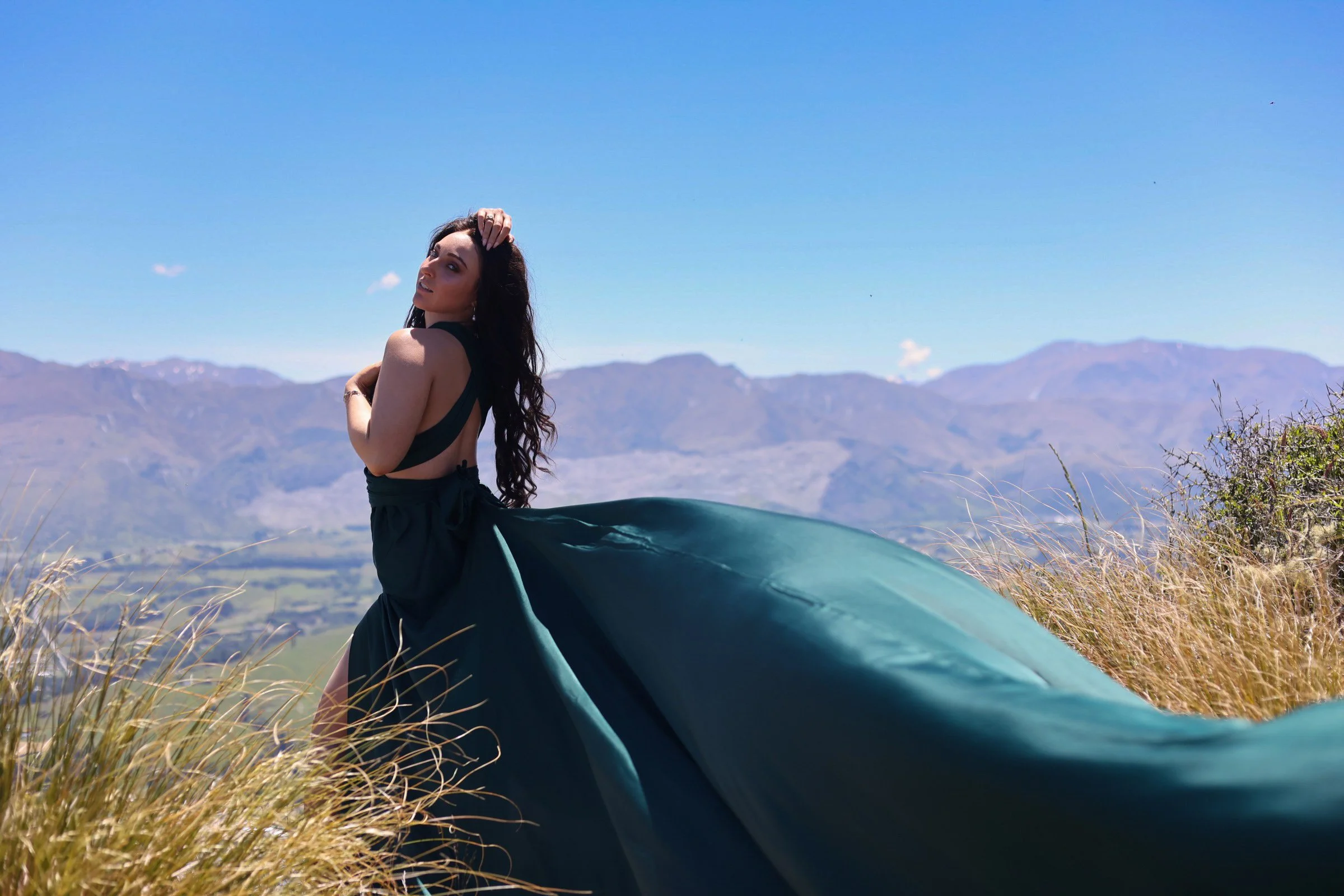 A woman with long dark hair wearing a black top and flowing skirt stands in a grassy field with mountains in the background under a clear blue sky.