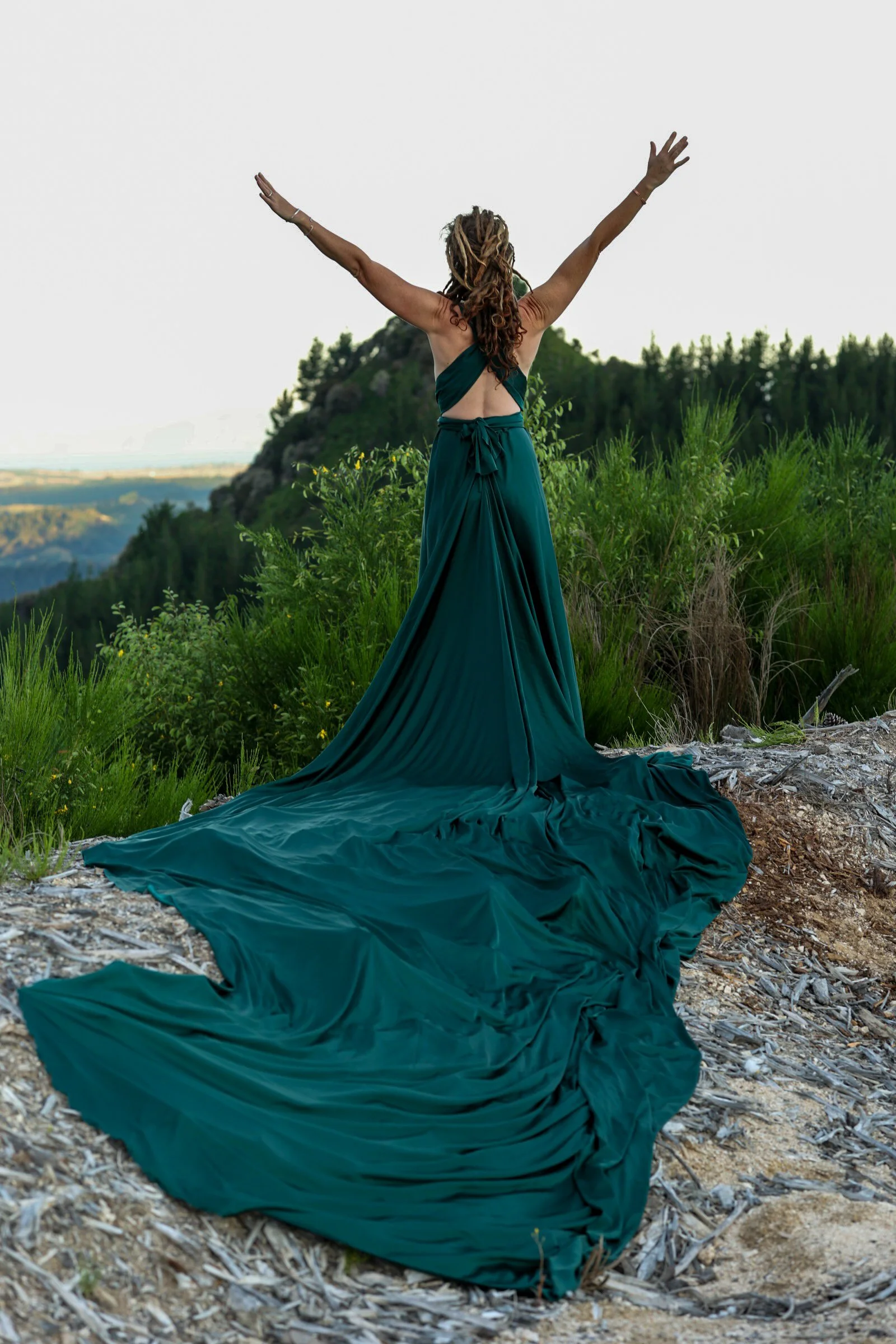 Woman with dreadlocks in a teal dress standing on a hilltop with arms raised, overlooking a landscape of trees and mountains in the distance.