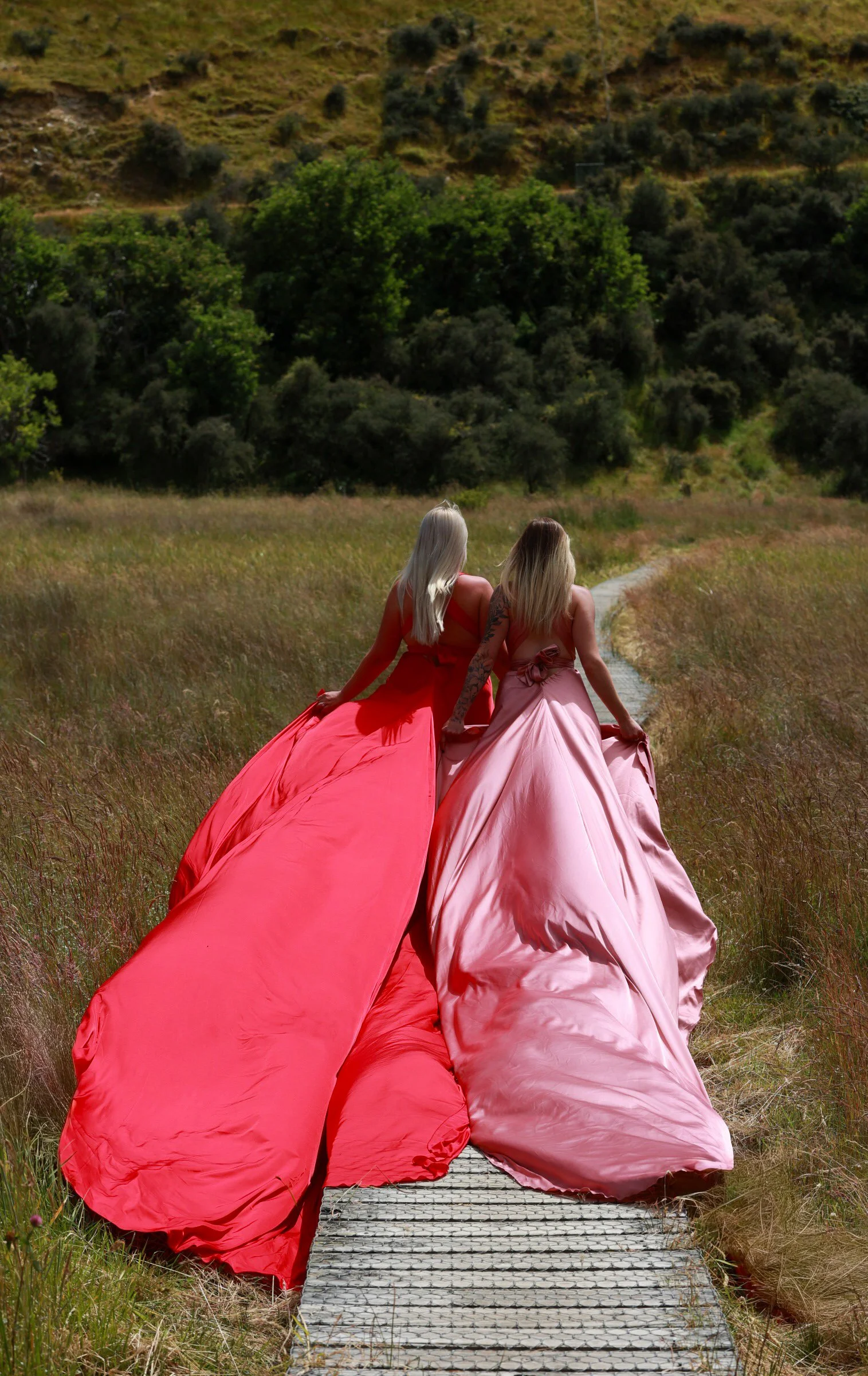 Two women in pink and red ball gowns walking away on a wooden path through a grassy field, with green trees and hills in the background.