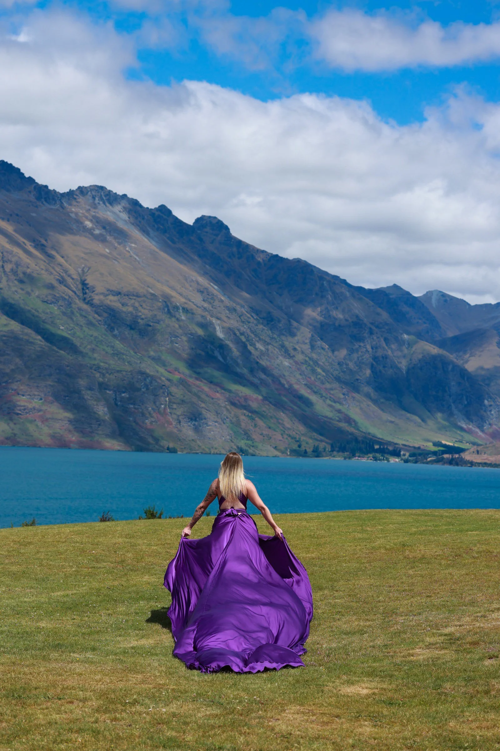 A woman with long blond hair in a flowing purple gown sitting on grass facing a lake and mountains in the distance under cloudy sky.