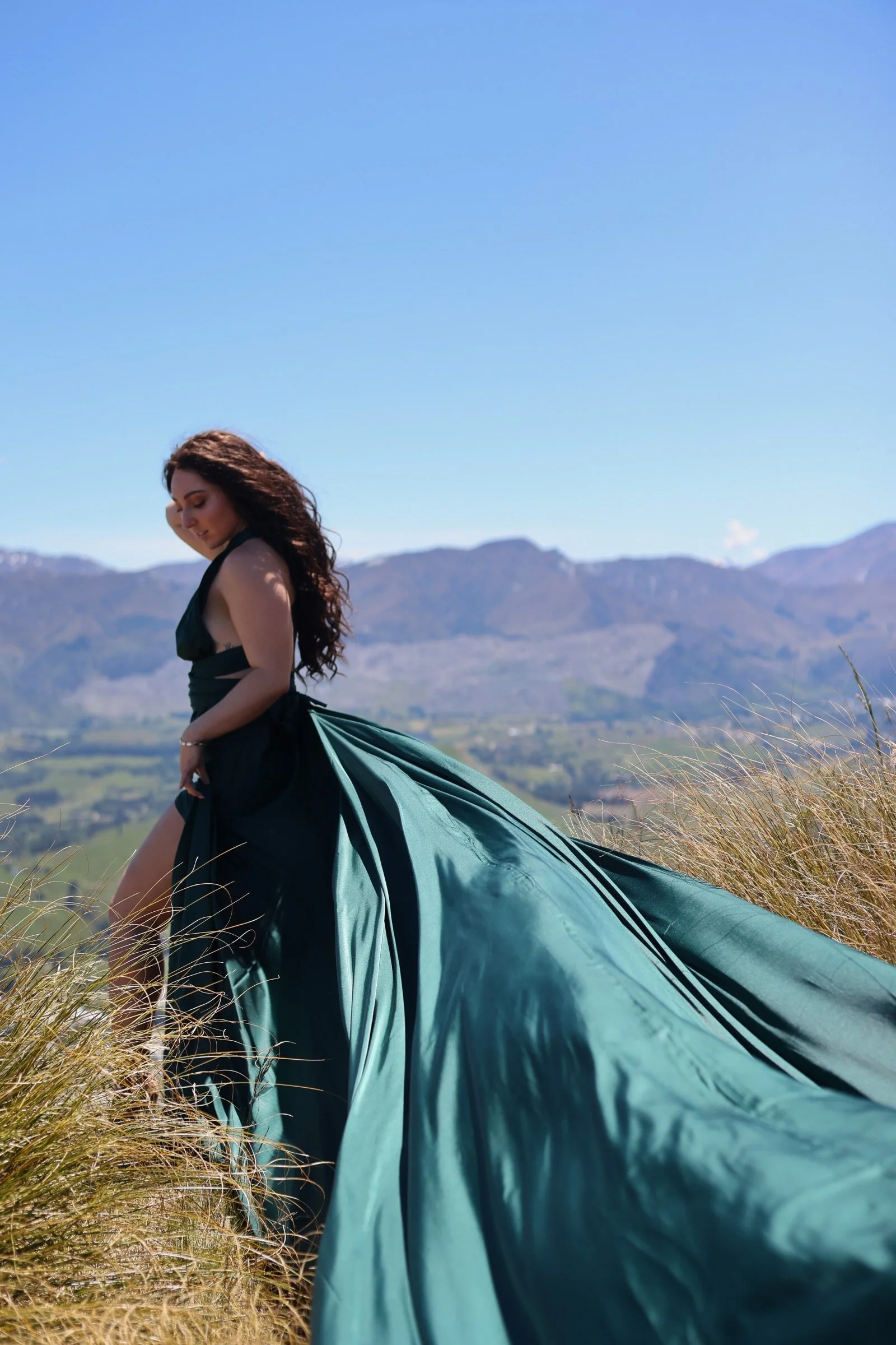 A woman with long, curly hair wearing a black dress stands in a grassy field with mountains in the background under a clear blue sky.