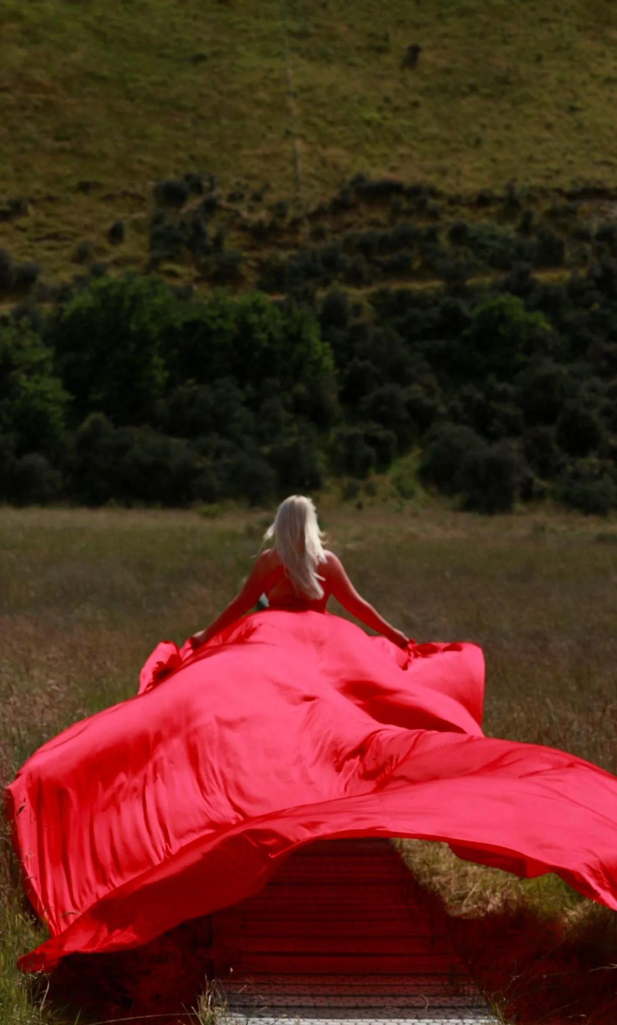 A woman with long blonde hair and a red dress sitting on a large red fabric, looking at a green hillside with trees and rocks.