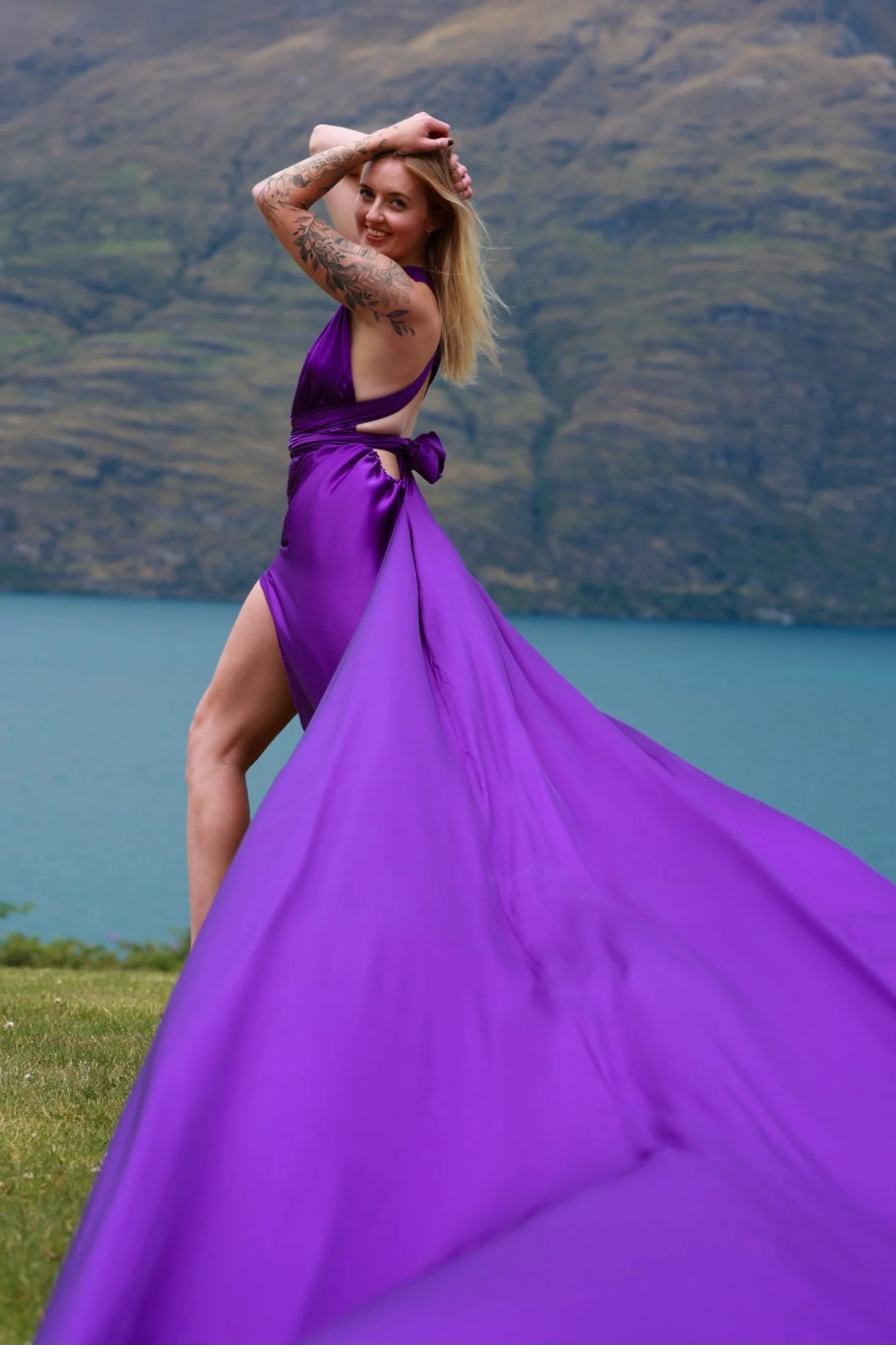 A woman in a vibrant purple dress with a high slit, standing outdoors near a lake with mountains in the background. She is smiling, with one hand on her head, revealing tattooed arms.