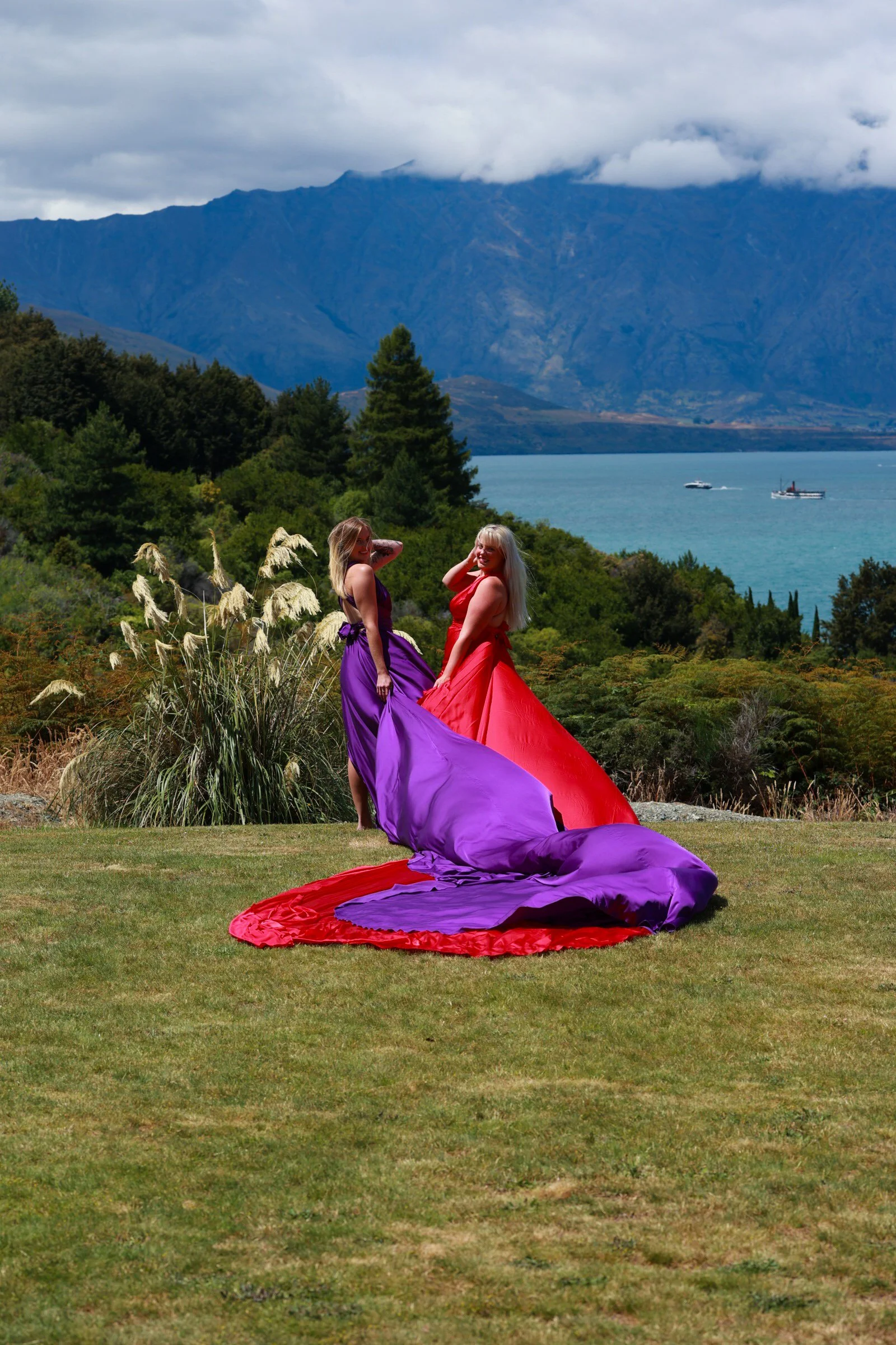 Two women wearing colorful dresses, one in purple and the other in red, standing outdoors on a grassy area with mountains, trees, and a lake in the background, and boats on the water.