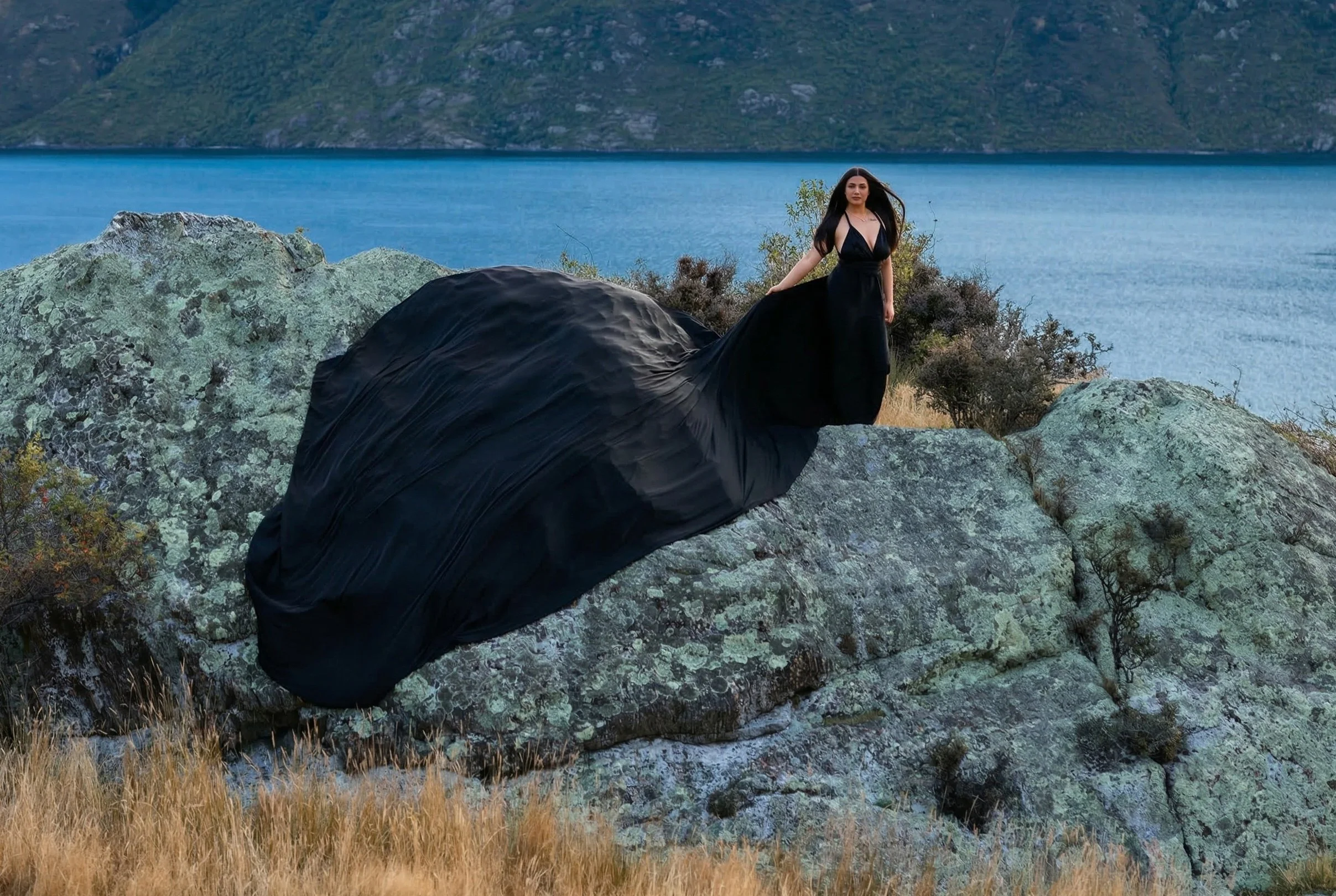 A woman in a long black dress stands on a rocky terrain near a body of water with mountains in the background.