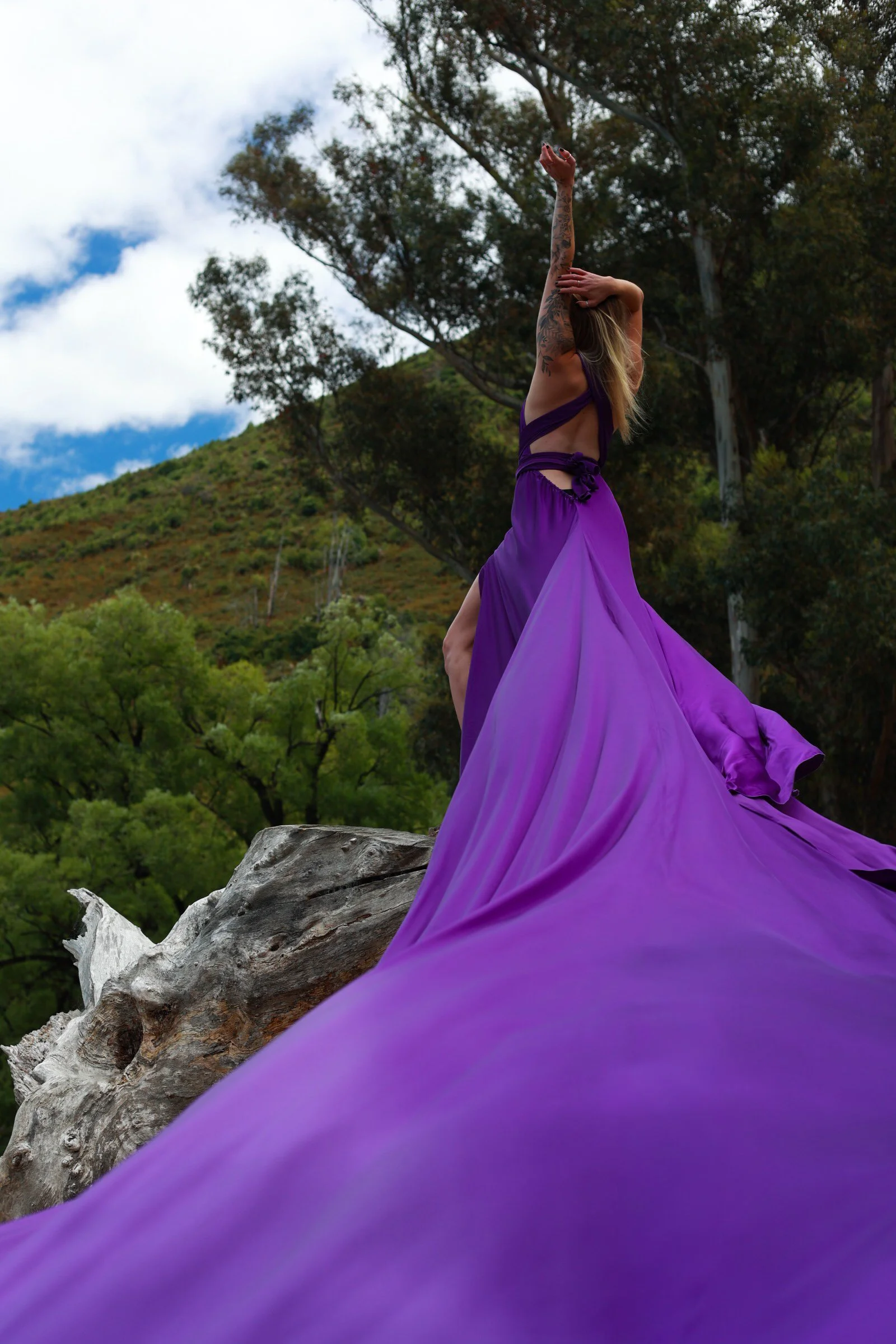 A woman with tattoos wearing a purple flowing dress, standing on a rock with her arms raised against a backdrop of trees, hills, and a partly cloudy sky.