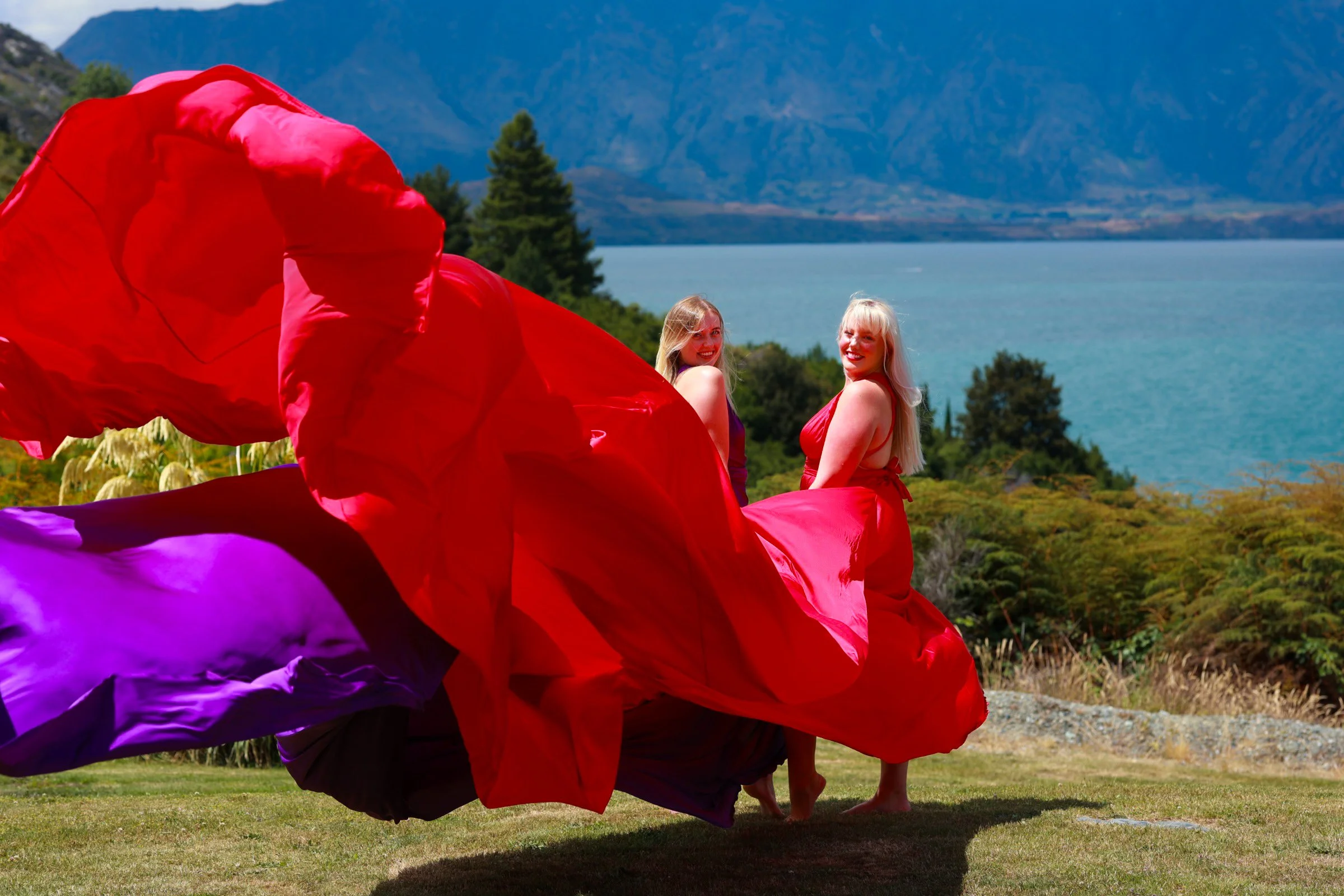 Two women with blonde hair standing outdoors near a body of water, wearing red dresses, with large flowing fabric in red, purple, and pink colors swirling around them, mountains and trees in the background.