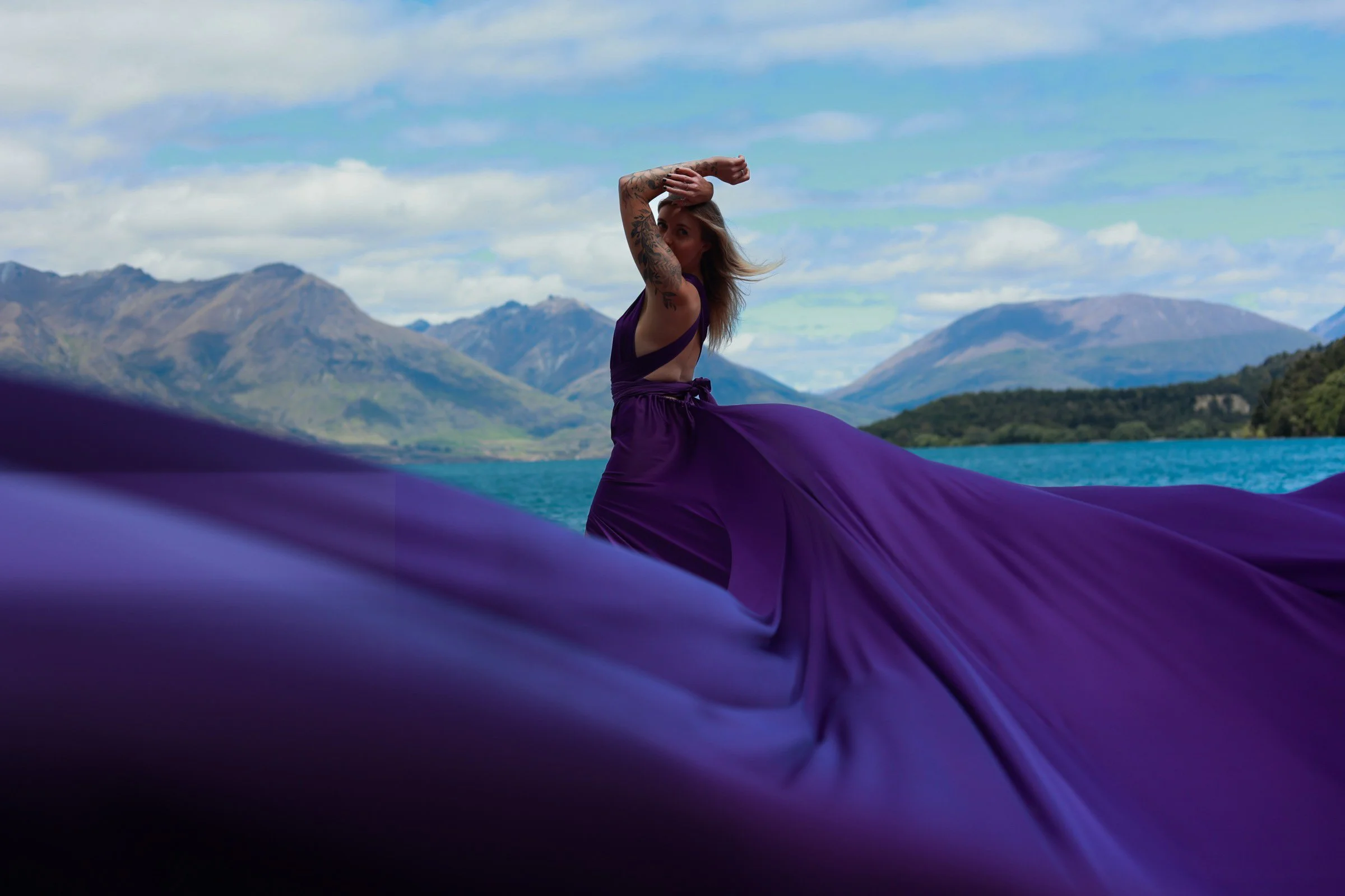 A woman wearing a purple dress is outdoors with mountains and a cloudy sky in the background. She is holding her arm above her head and appears to be dancing or performing.
