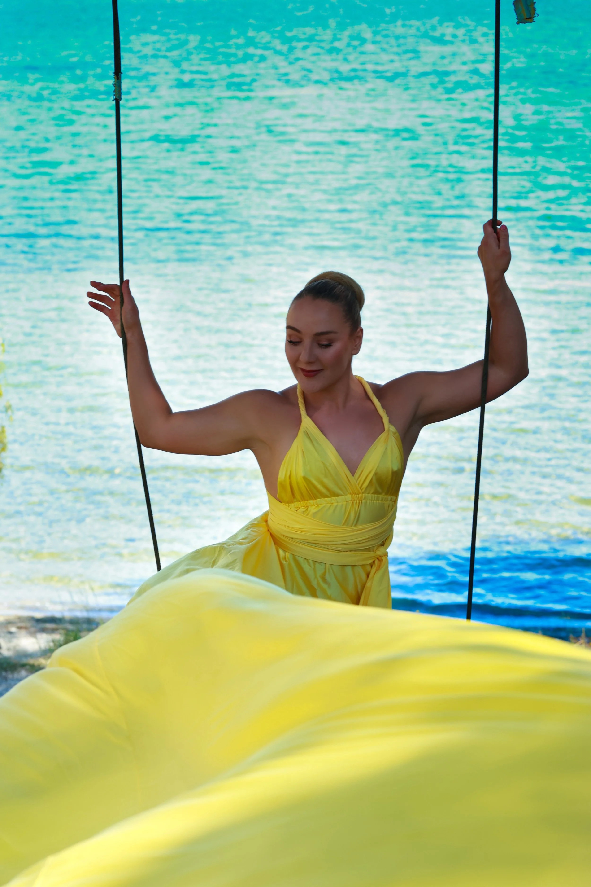 A woman in a yellow dress sitting on a swing by a lake, with her eyes closed and a gentle smile.