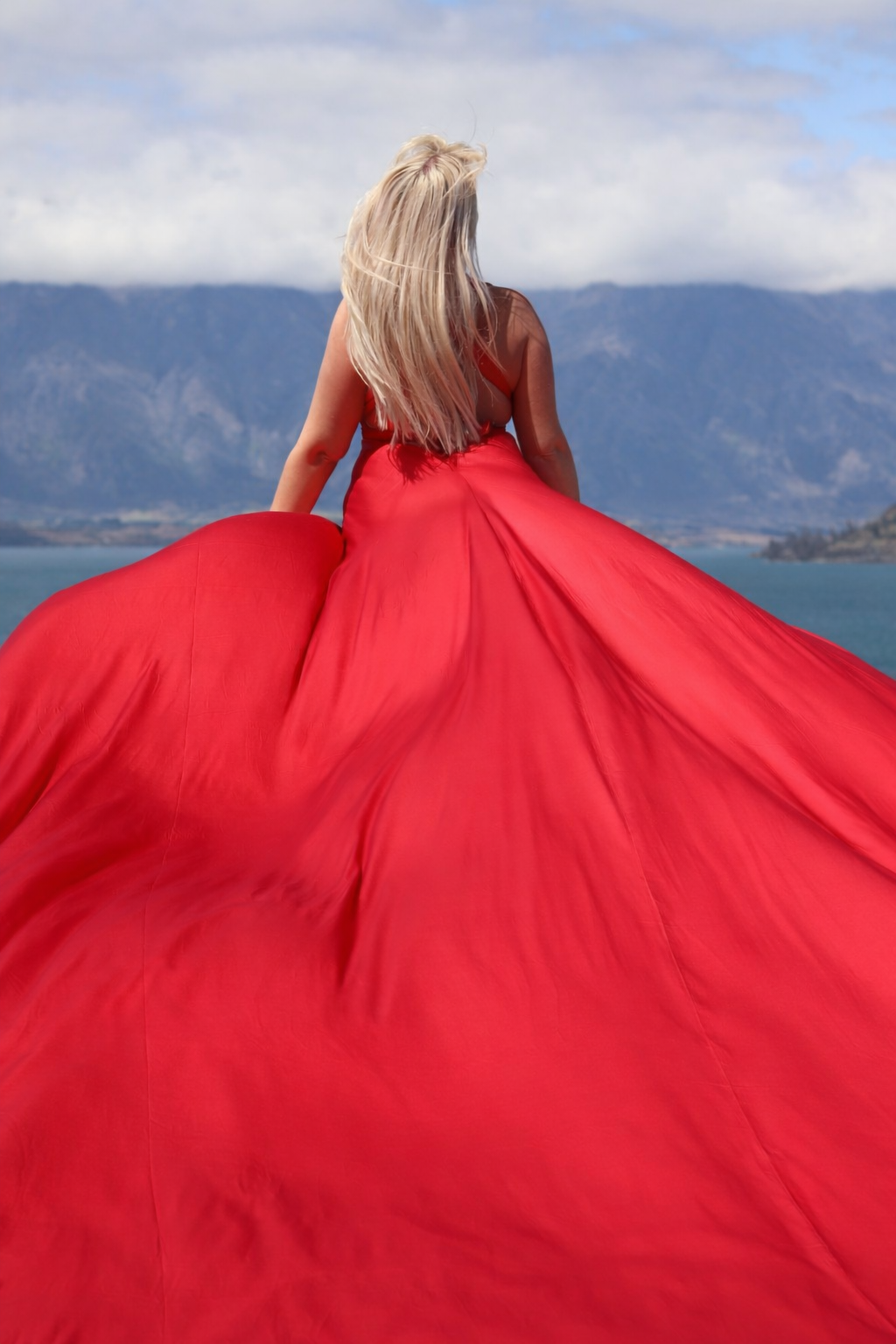 A woman with long blonde hair wearing a flowing red gown standing on a scenic landscape with mountains, lake, and cloudy sky in the background.