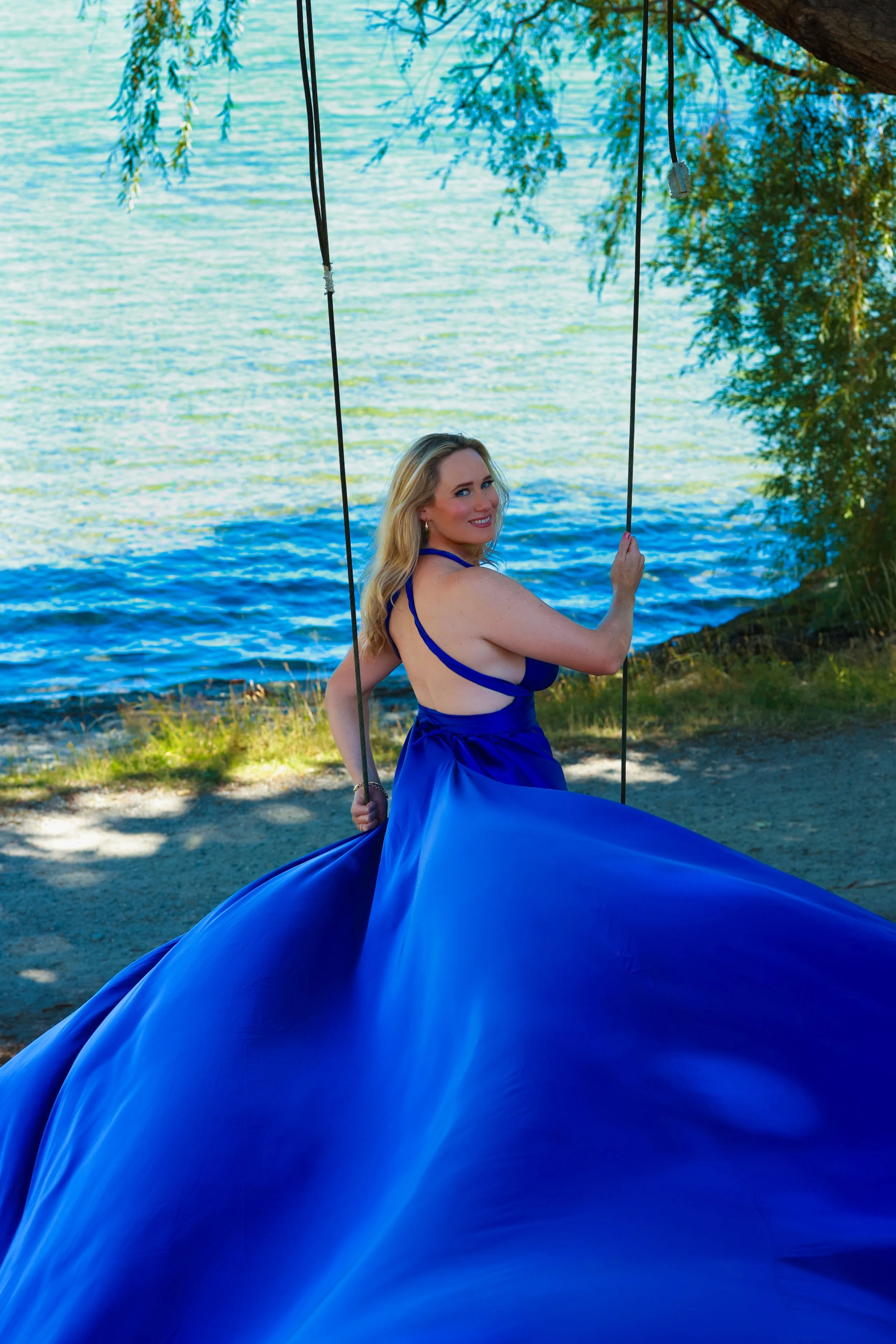 A woman in a blue dress sitting on a swing by the water, with trees and water in the background.