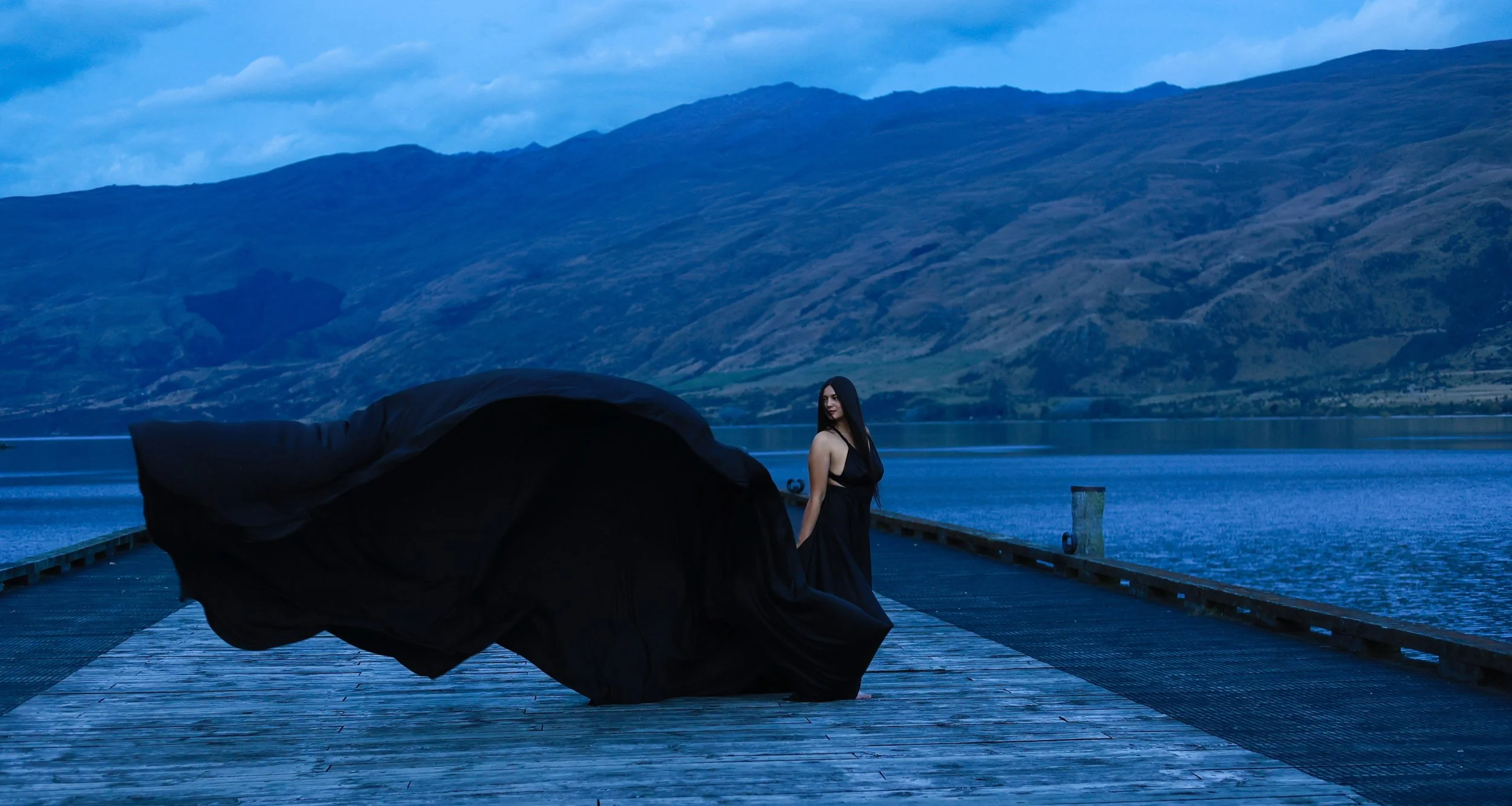 A woman in a long black dress standing on a wooden pier by a lake, with mountains in the background, during dusk.