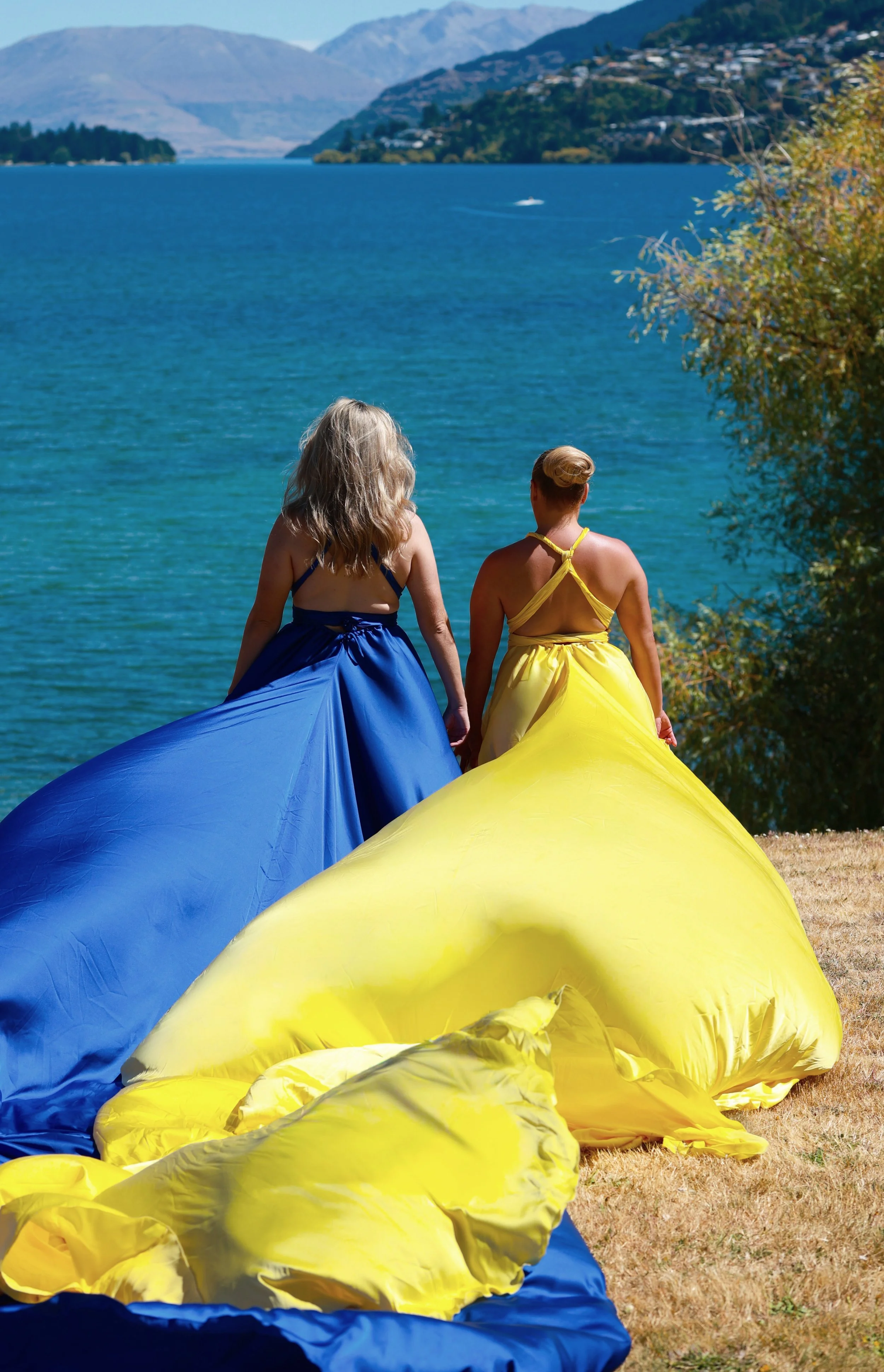 Two women in colorful dresses, one in blue and the other in yellow, walk away from the camera towards a lake with mountains in the background, carrying inflatables.