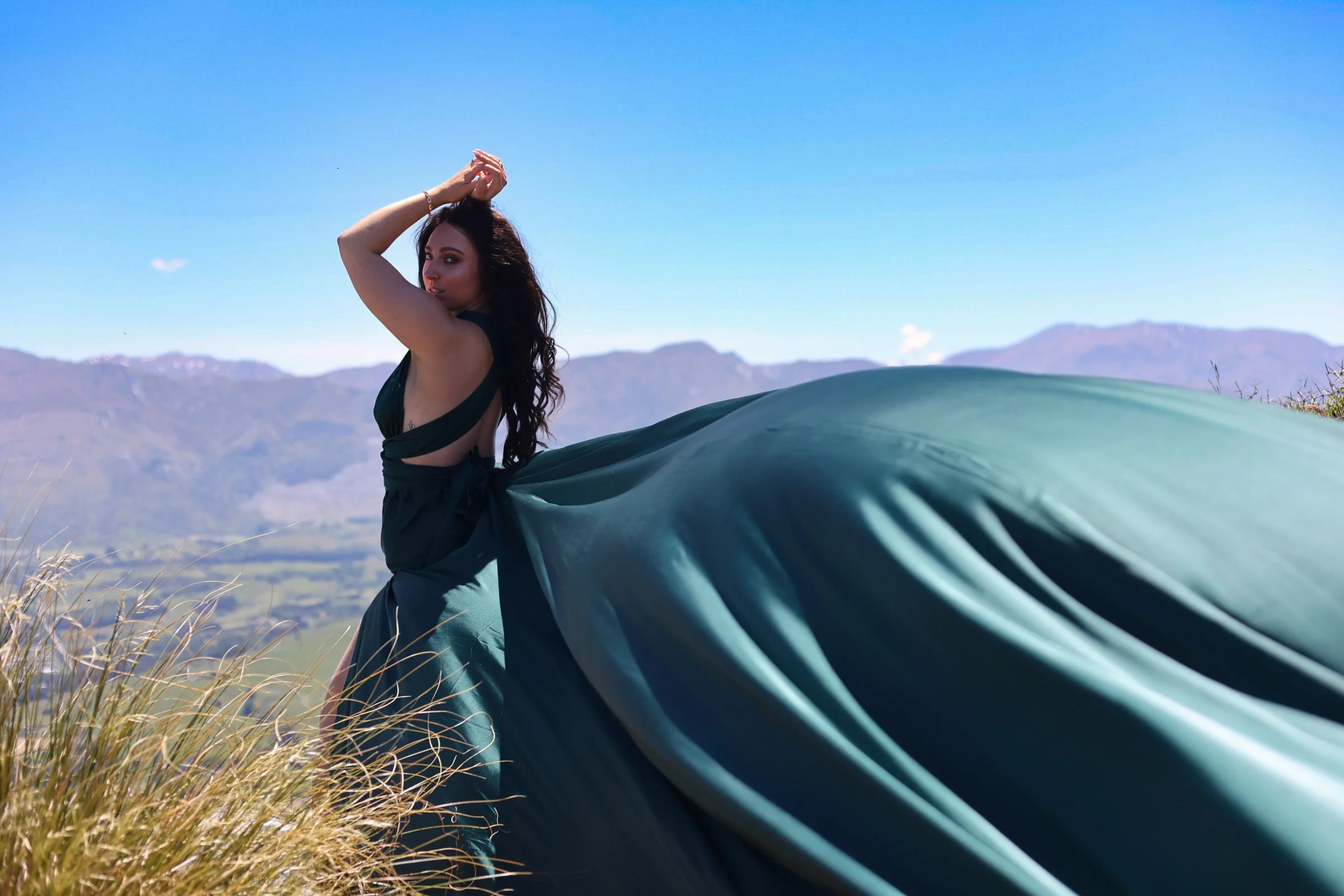 A woman with long dark hair standing outdoors on a hilltop, wearing a black top and flowing teal skirt, with mountains and a clear blue sky in the background.