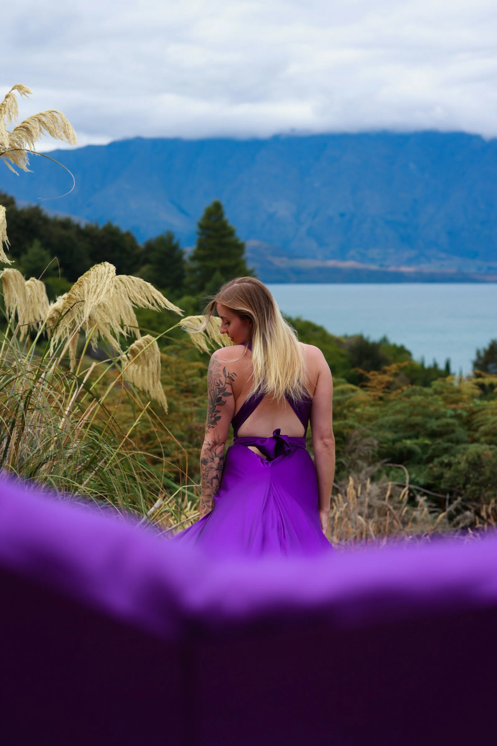 A woman with blonde hair and tattoos wearing a purple dress standing outdoors with a mountain and lake in the background, surrounded by tall grasses.