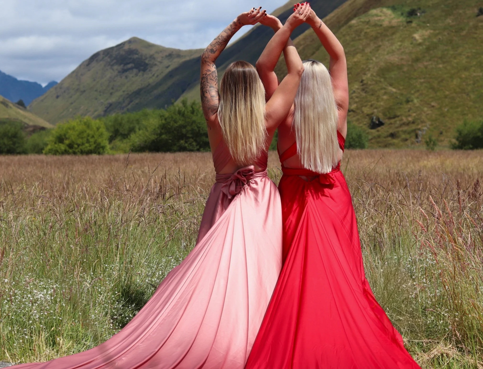 Two women with long blonde hair in red dresses standing in a grassy field with mountains in the background. They are facing away from the camera and holding hands above their heads, forming a heart shape. One woman has tattoos on her arms.