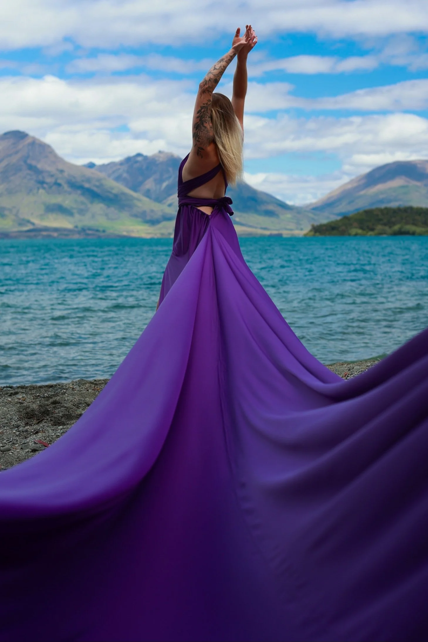 A woman in a purple dress standing on a beach with mountain scenery in the background, raising her arms above her head.