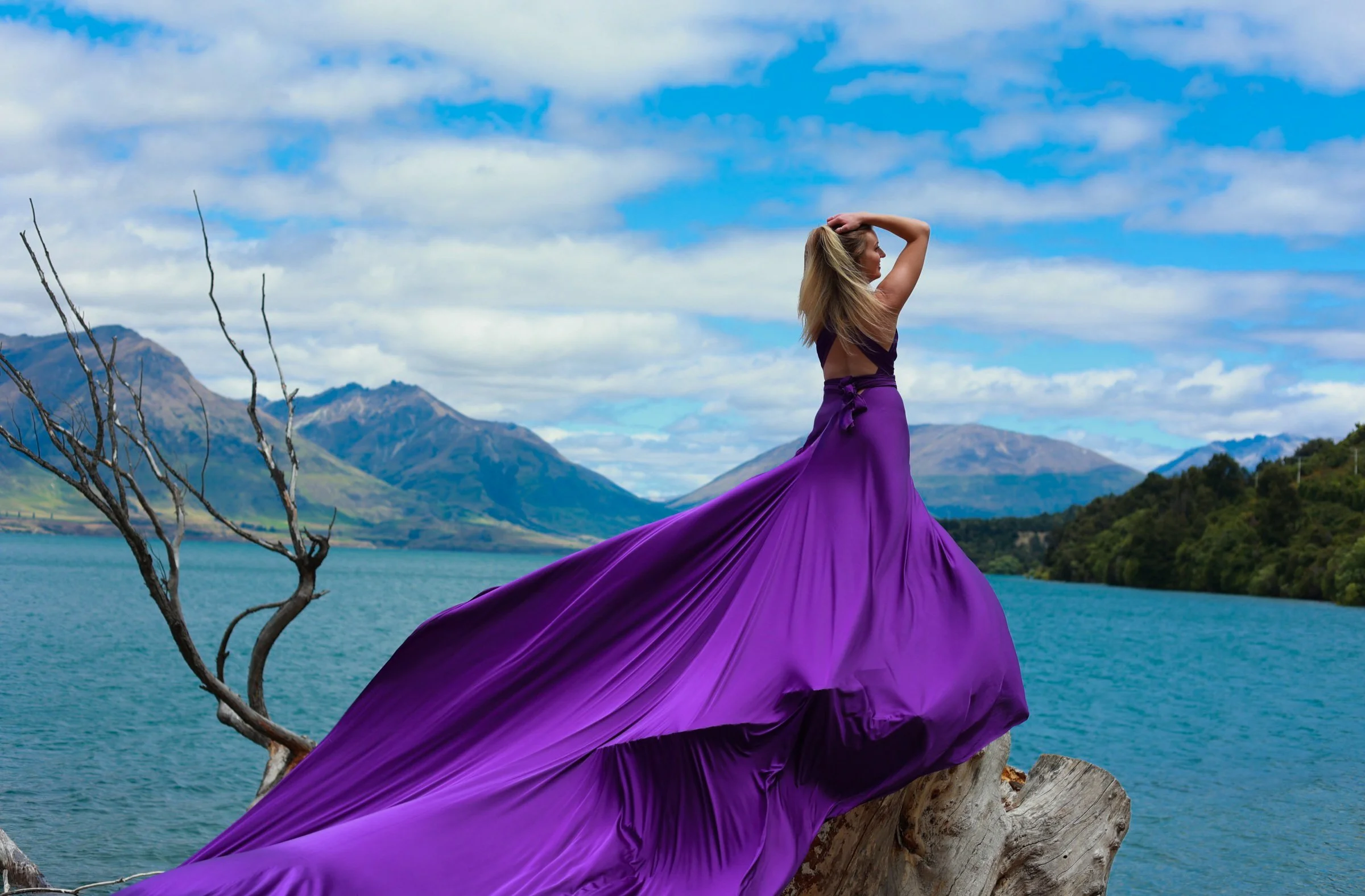 A woman in a flowing purple dress standing on a tree trunk near a body of water with mountains in the background and a partly cloudy sky.