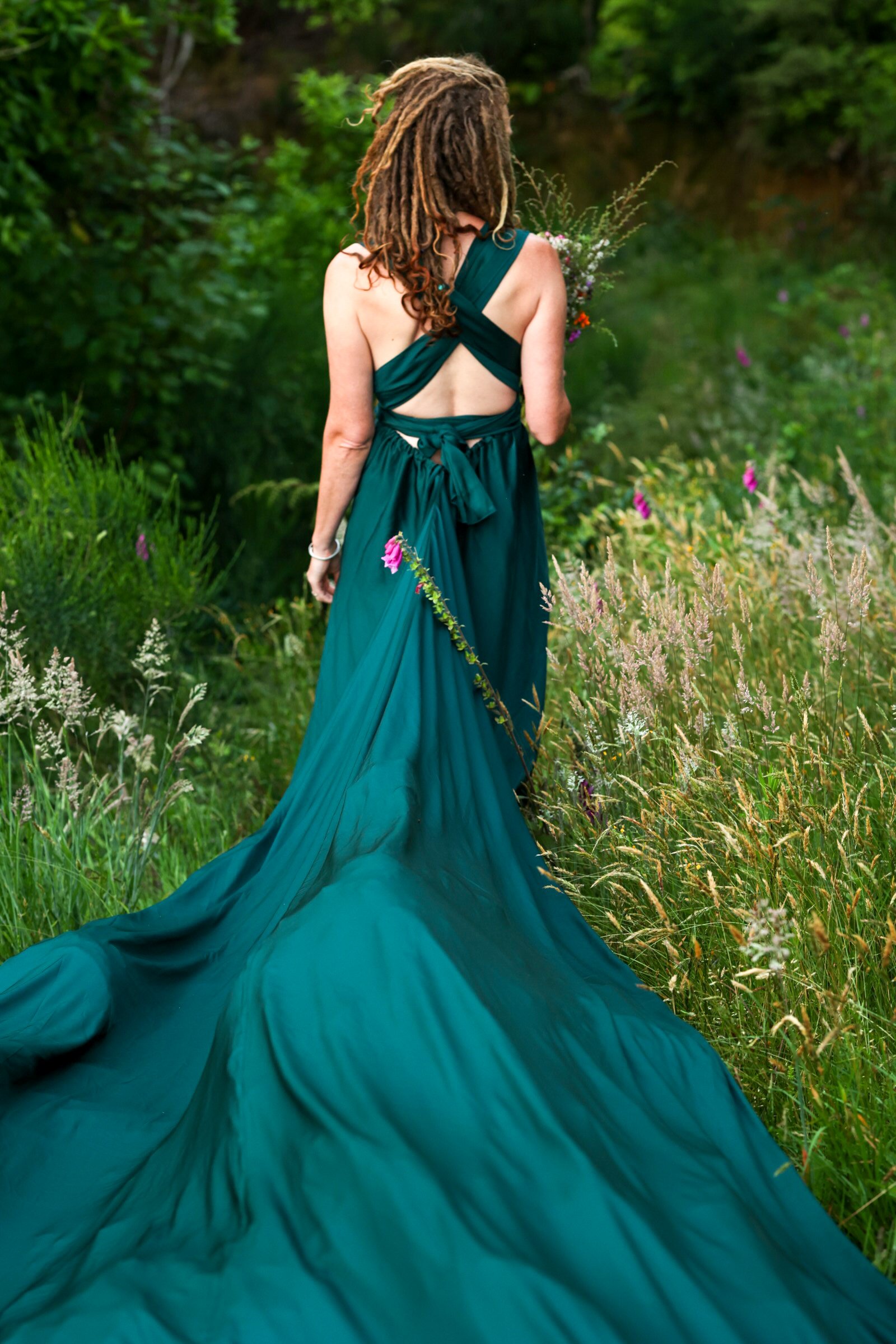 Back of a woman with dreadlocks wearing a dark green flowing dress, standing in a lush garden with various green plants and flowers.