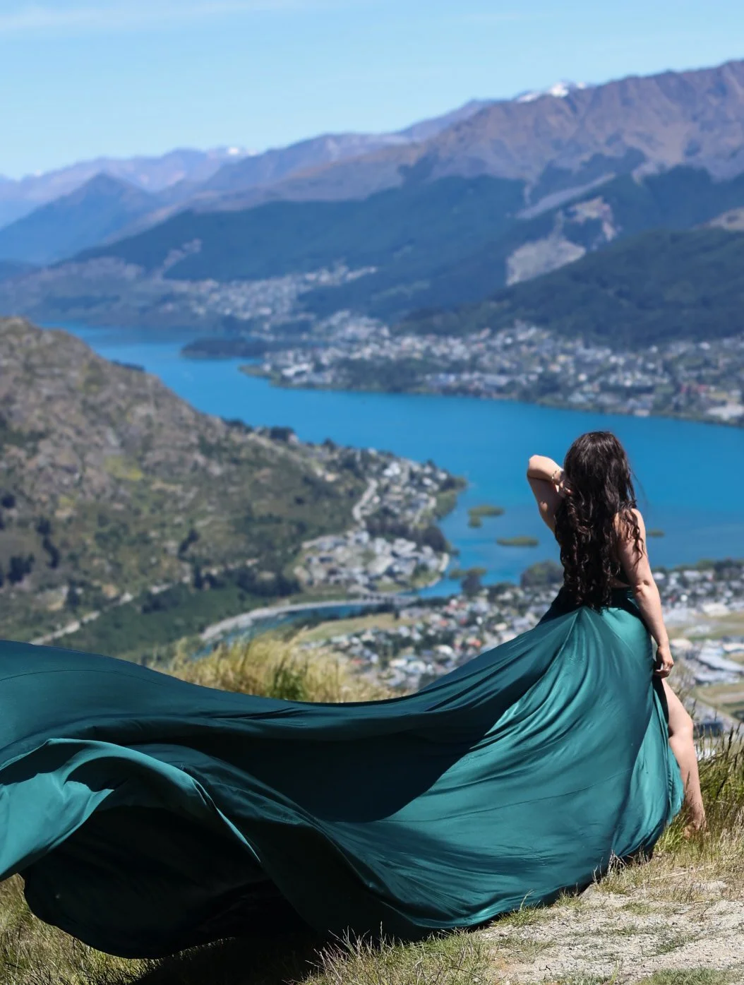 A woman wearing a long teal dress sitting on a grassy hill overlooking a river, mountains, and a town in the distance.