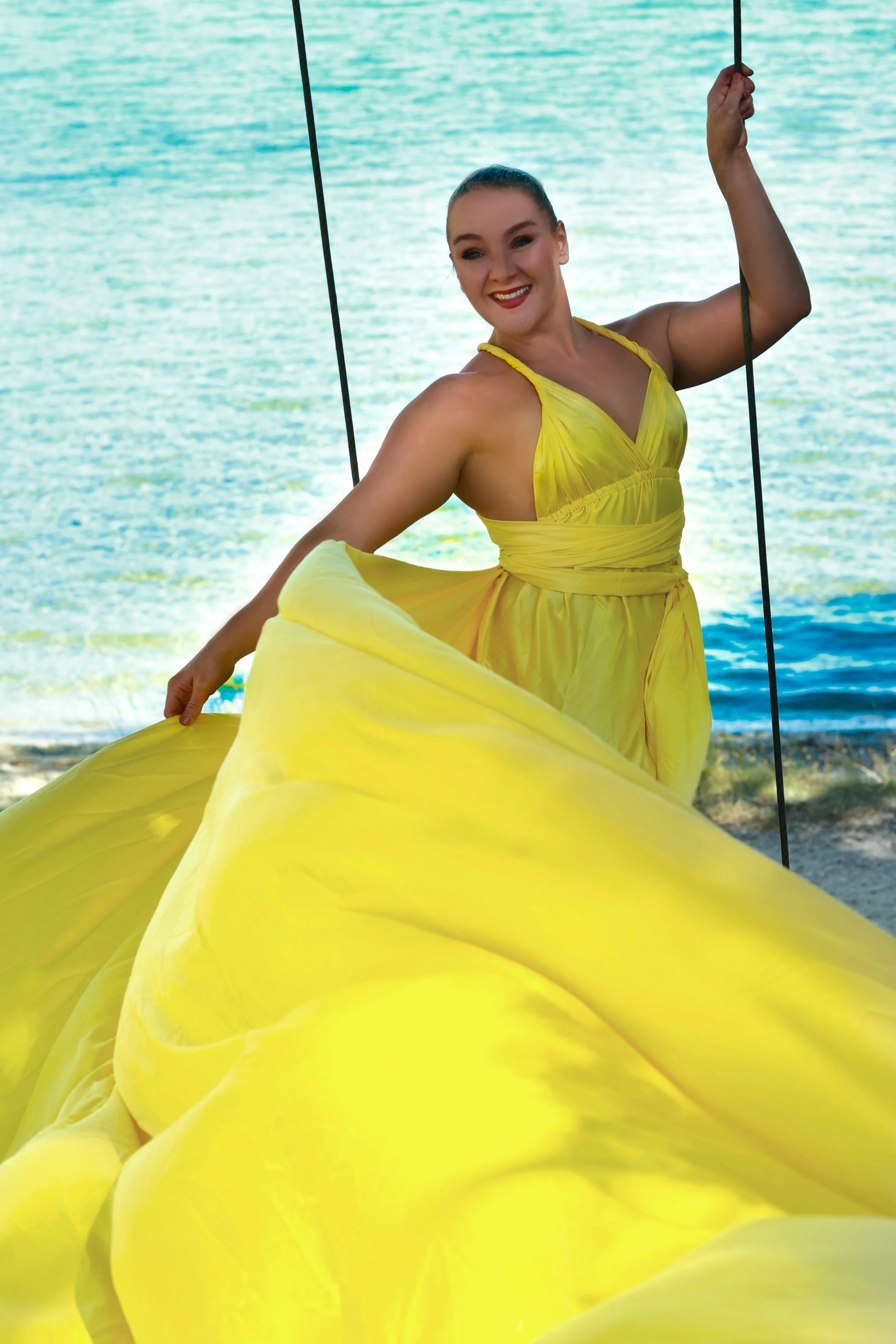 A woman in a yellow dress smiling and holding onto swings in front of a body of water, with trees and a shoreline in the background.