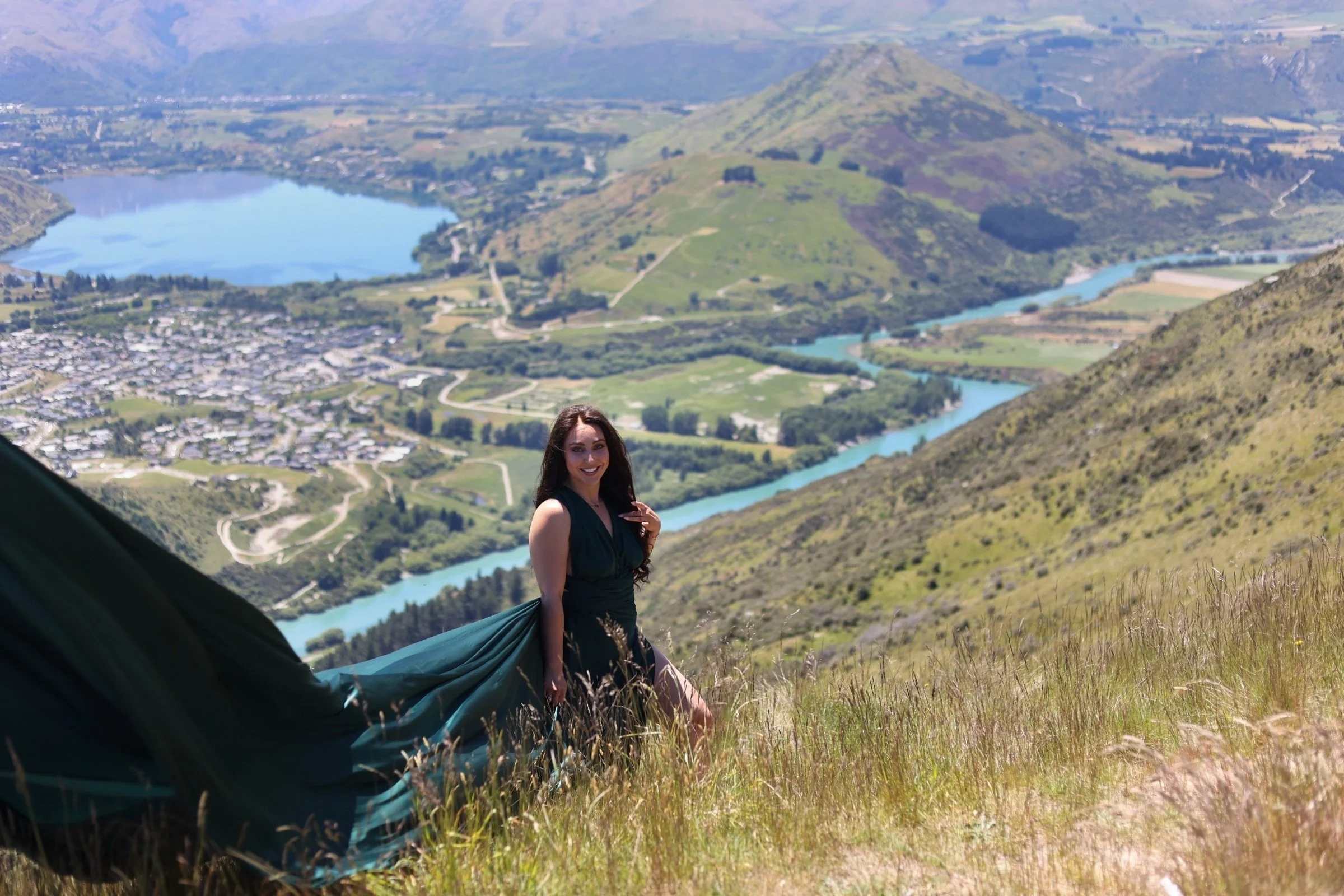A woman with long dark hair wearing a teal dress, sitting in a grassy hillside with mountains, a lake, and a river in the background.