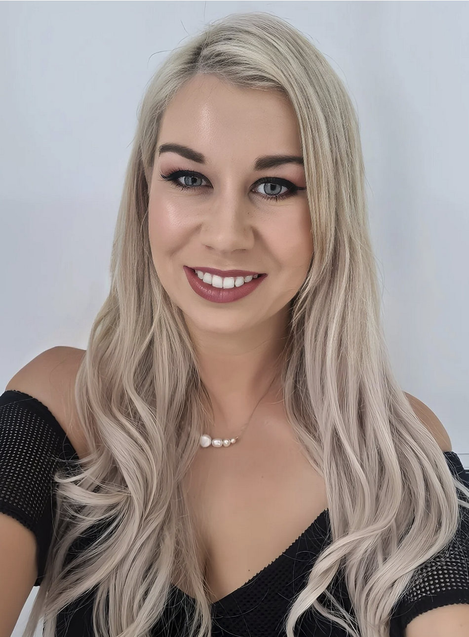 A young woman with long, wavy, platinum blonde hair, blue eyes, and light makeup, wearing a black off-shoulder top and a pearl necklace, smiling at the camera against a plain white background.