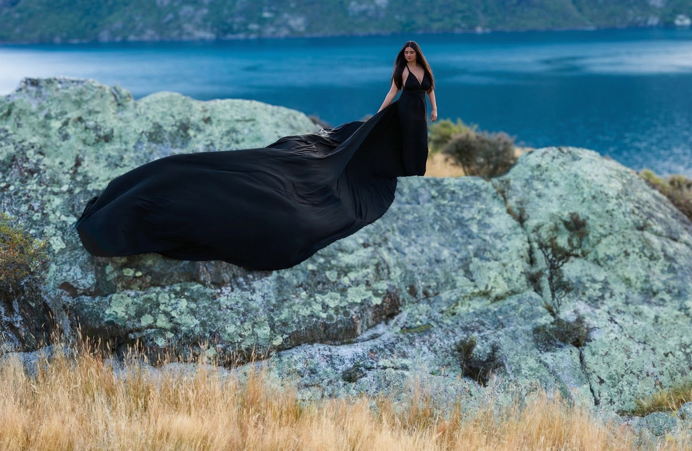 A woman in a long black evening gown stands on rocky terrain with a body of water and hills in the background.