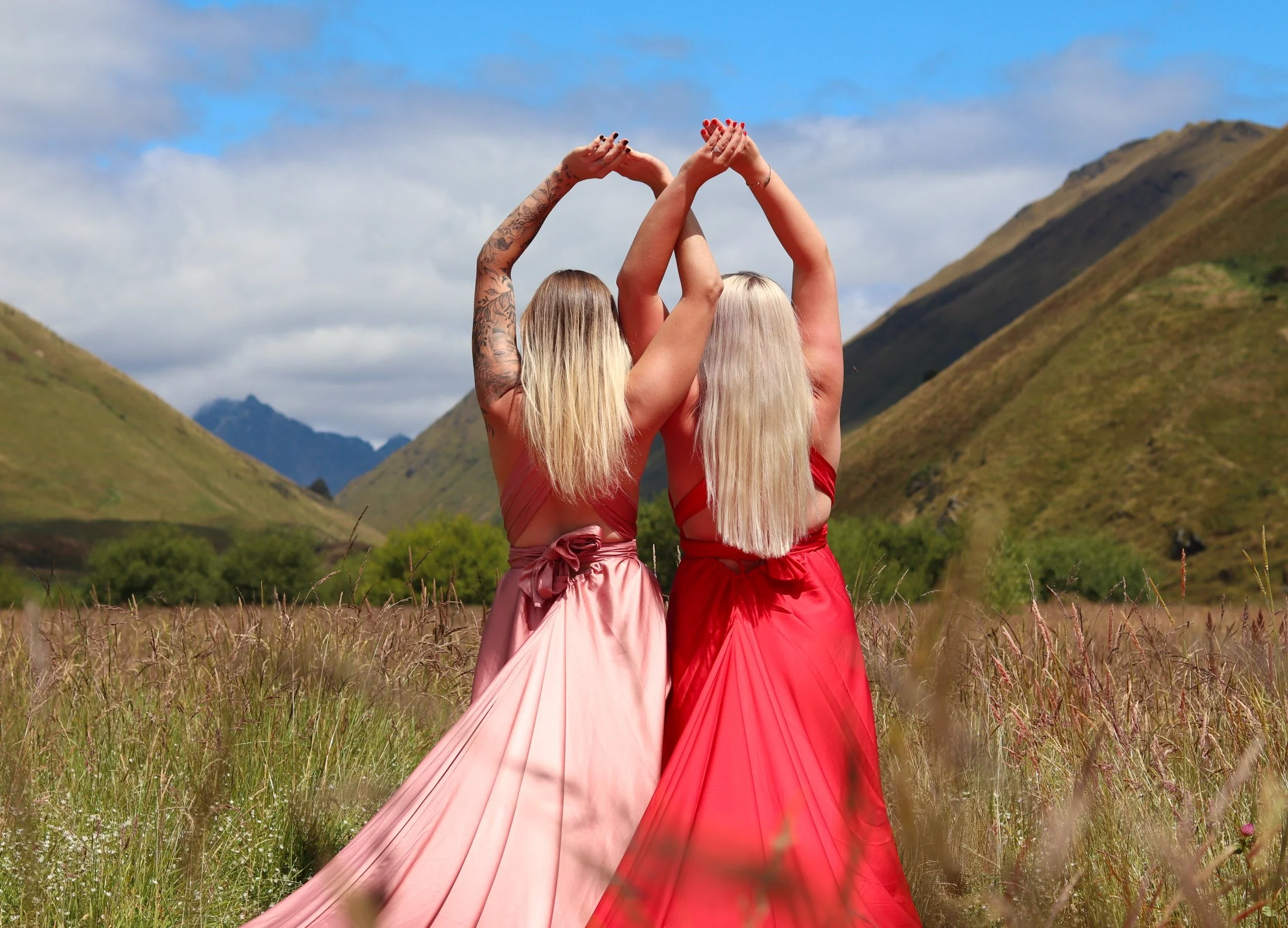 Two women in colorful dresses standing outdoors in a field with mountains in the background, raising their hands together.