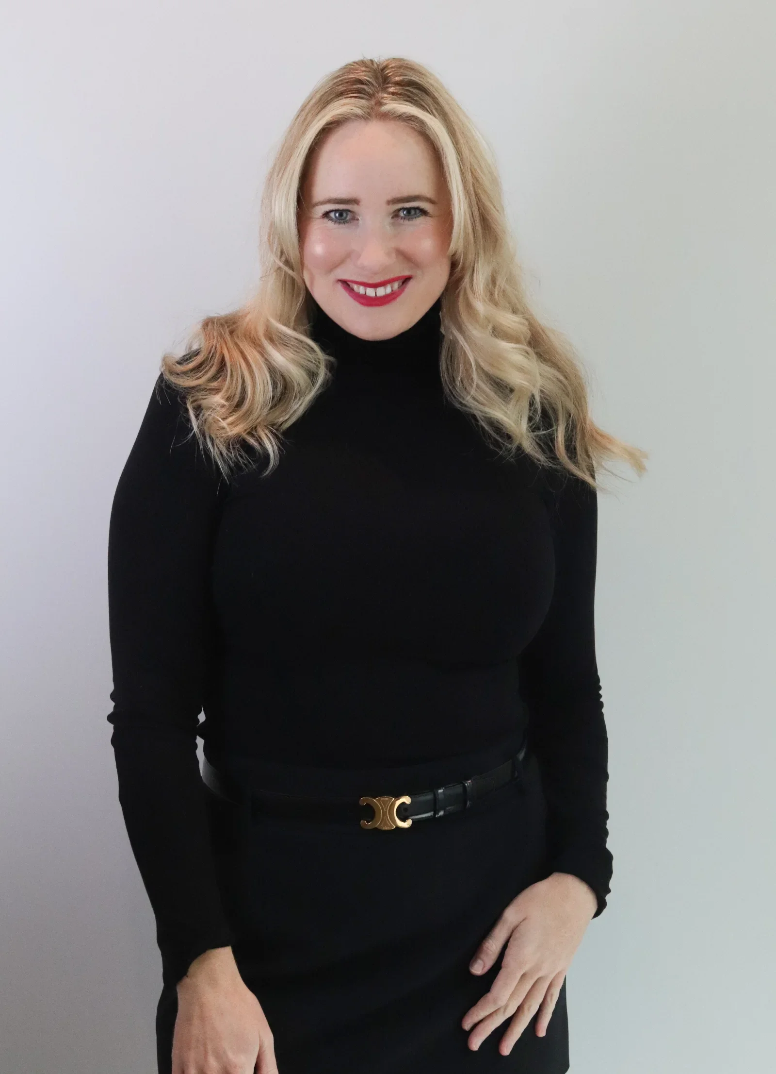 A woman with blonde hair wearing a black turtleneck and black skirt standing against a plain white wall.