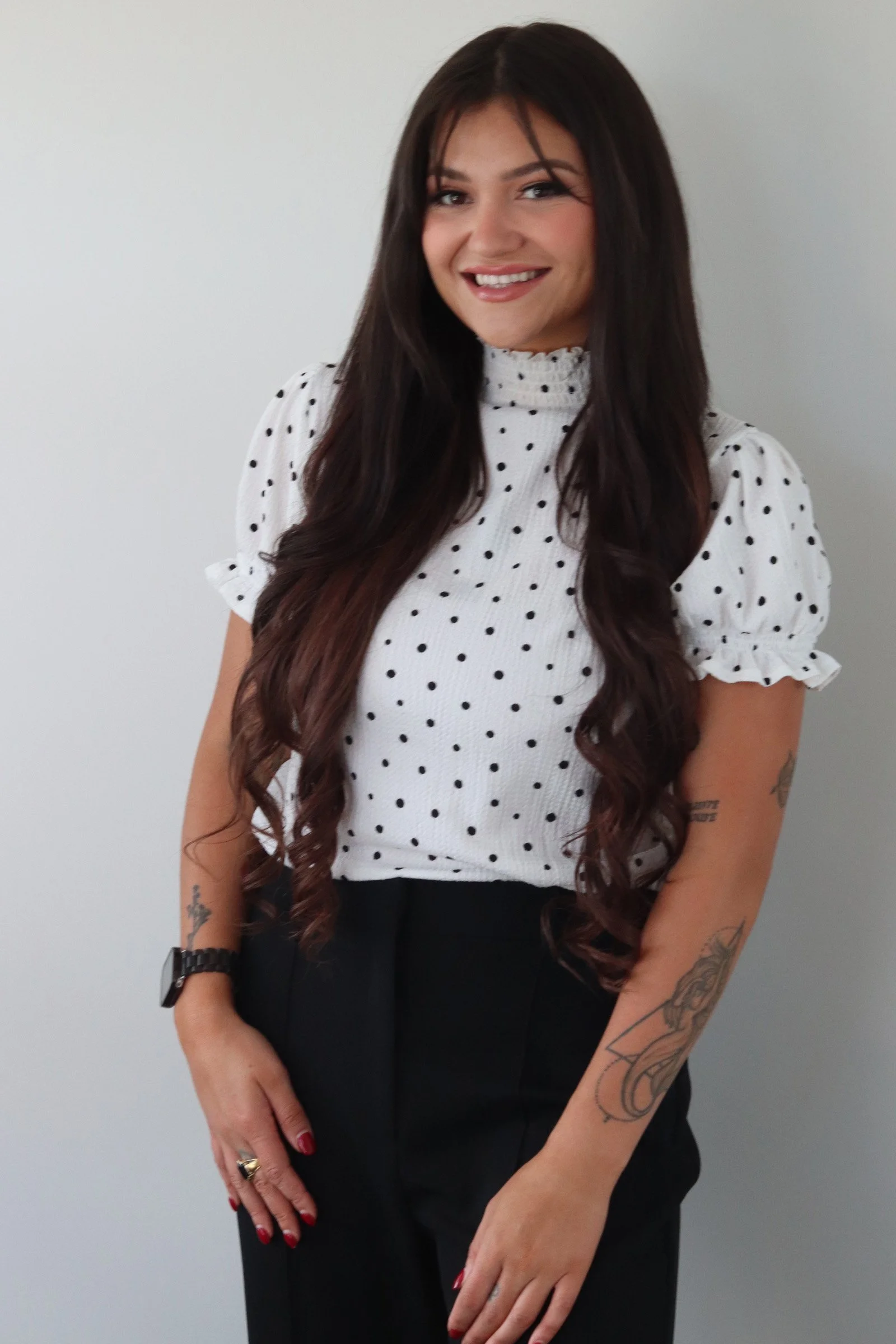A young woman with long dark hair, smiling, wearing a white top with black polka dots and black pants, standing against a plain gray background.