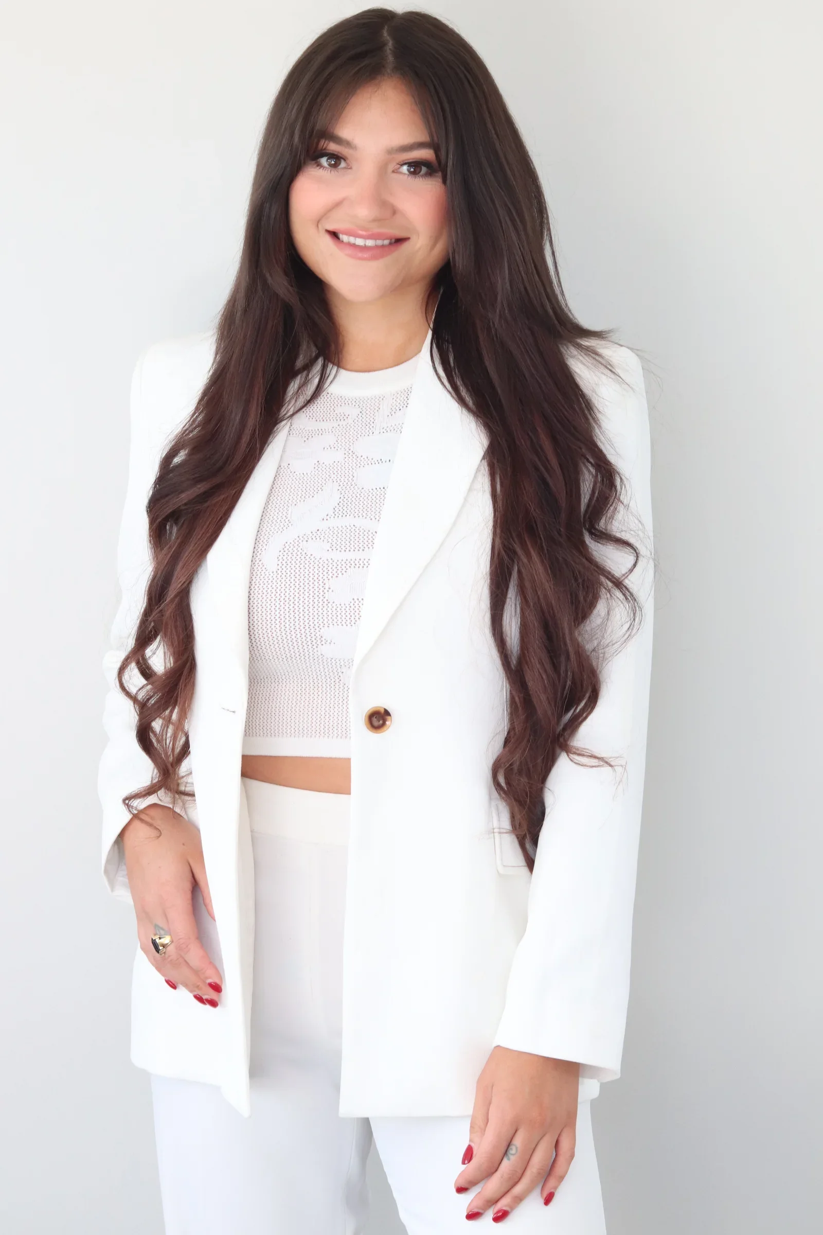 A woman with long dark brown hair, wearing a white suit and a white lace crop top, smiling while standing against a plain white background.