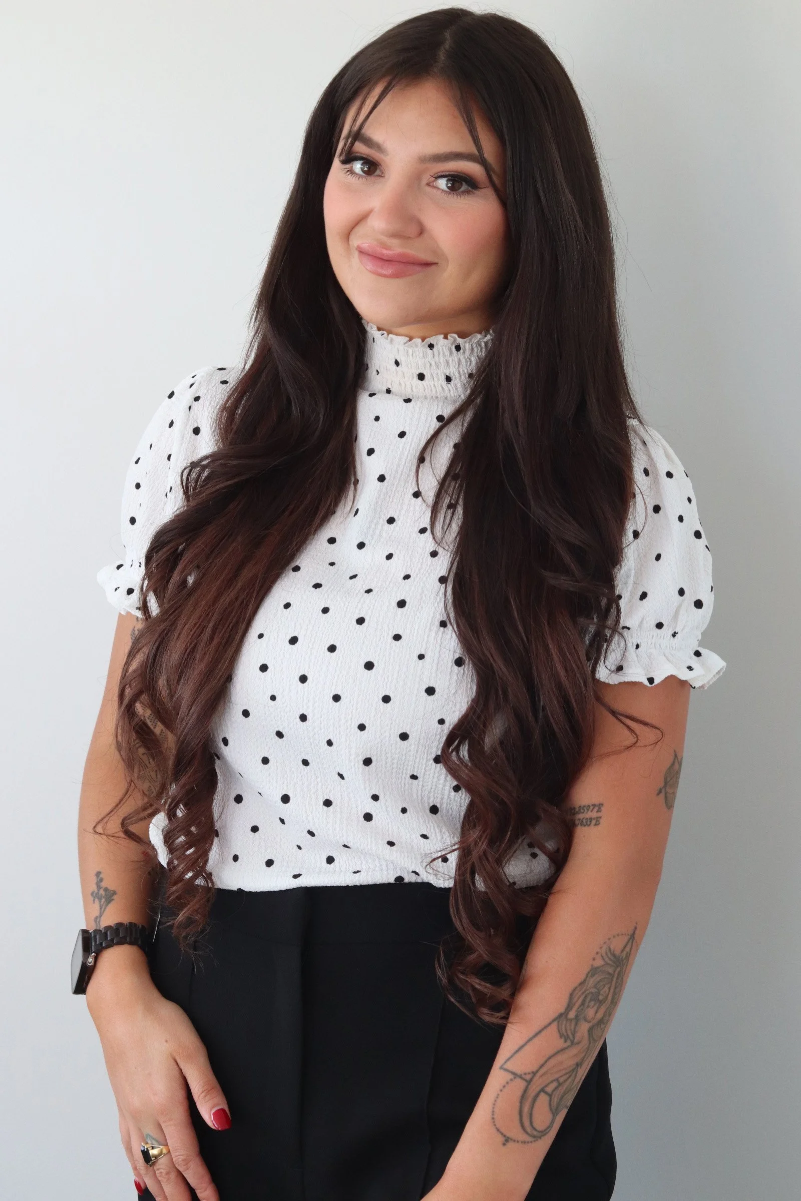 A woman with long, dark curly hair wearing a white polka dot blouse and black pants, smiling with her head slightly tilted, standing against a plain light-colored wall.