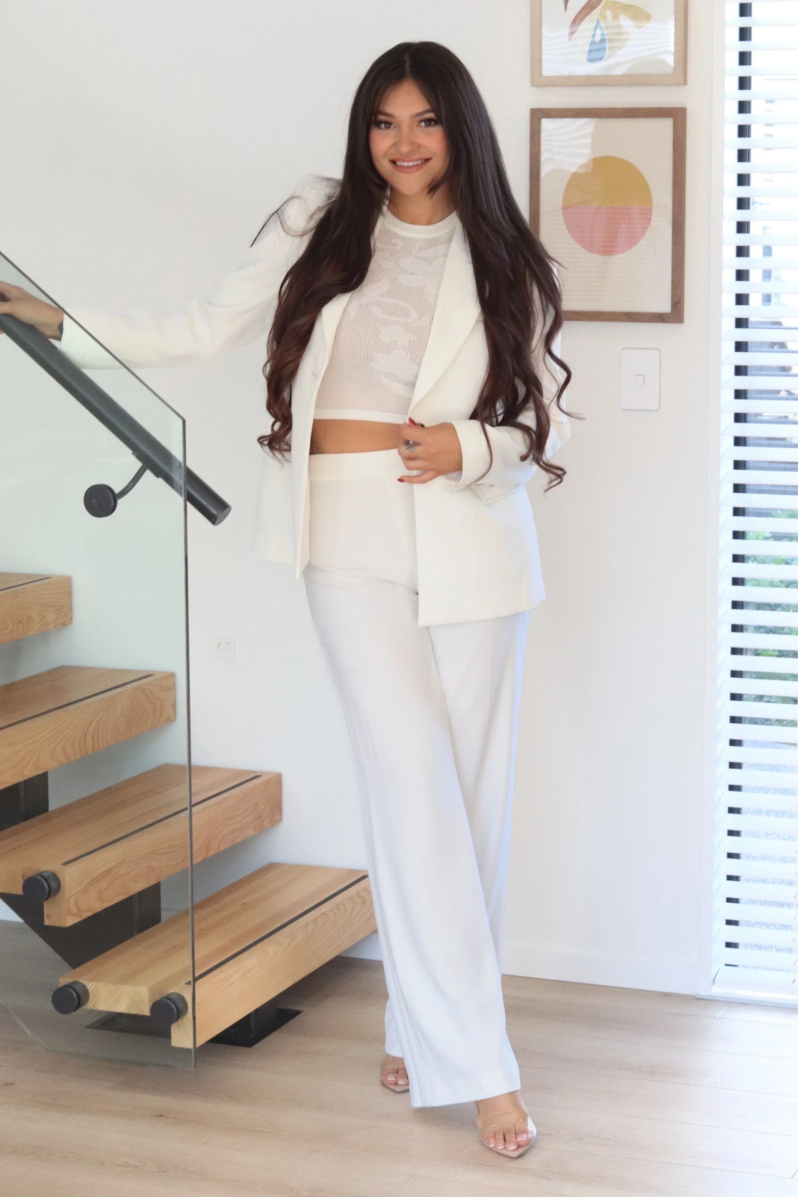A woman with long, wavy dark hair poses in a white suit, standing near a staircase with wooden steps and glass railing, inside a modern room with white walls and framed abstract art.