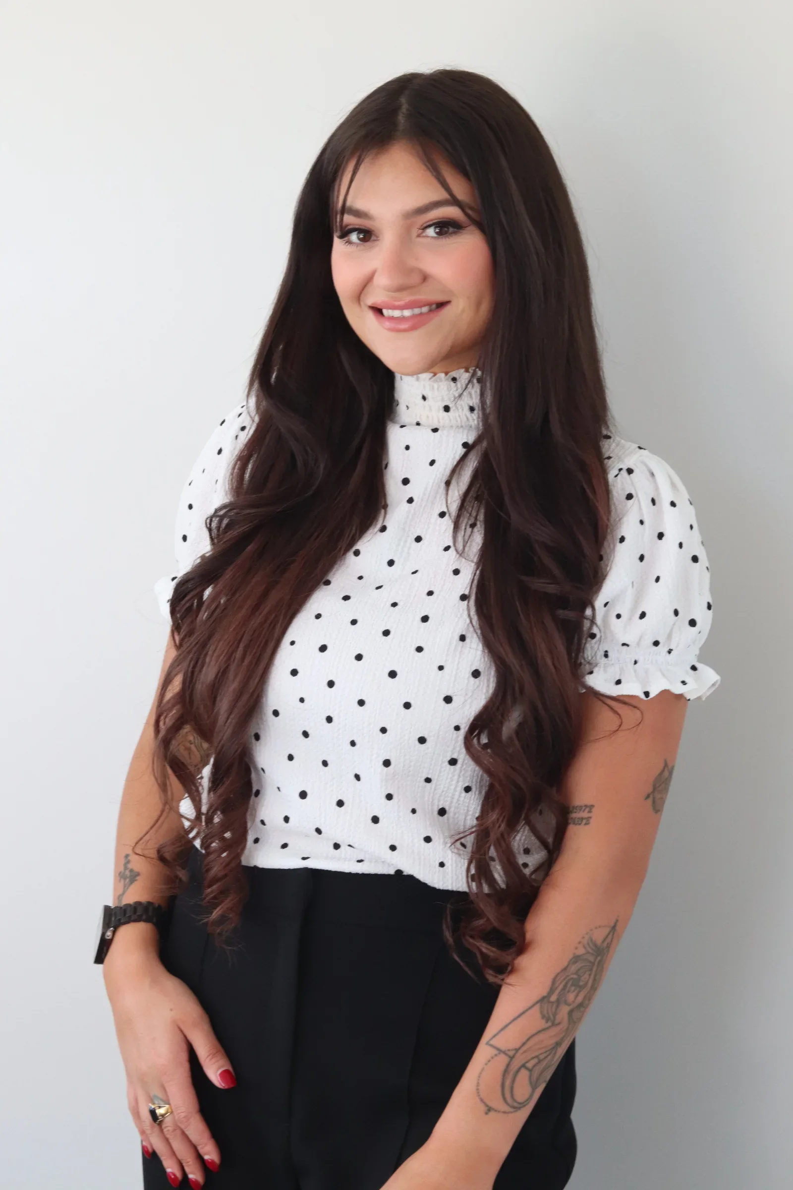 A young woman with long, dark hair and tattoos on her arms, smiling, wearing a white polka dot blouse and black pants, standing against a white background.