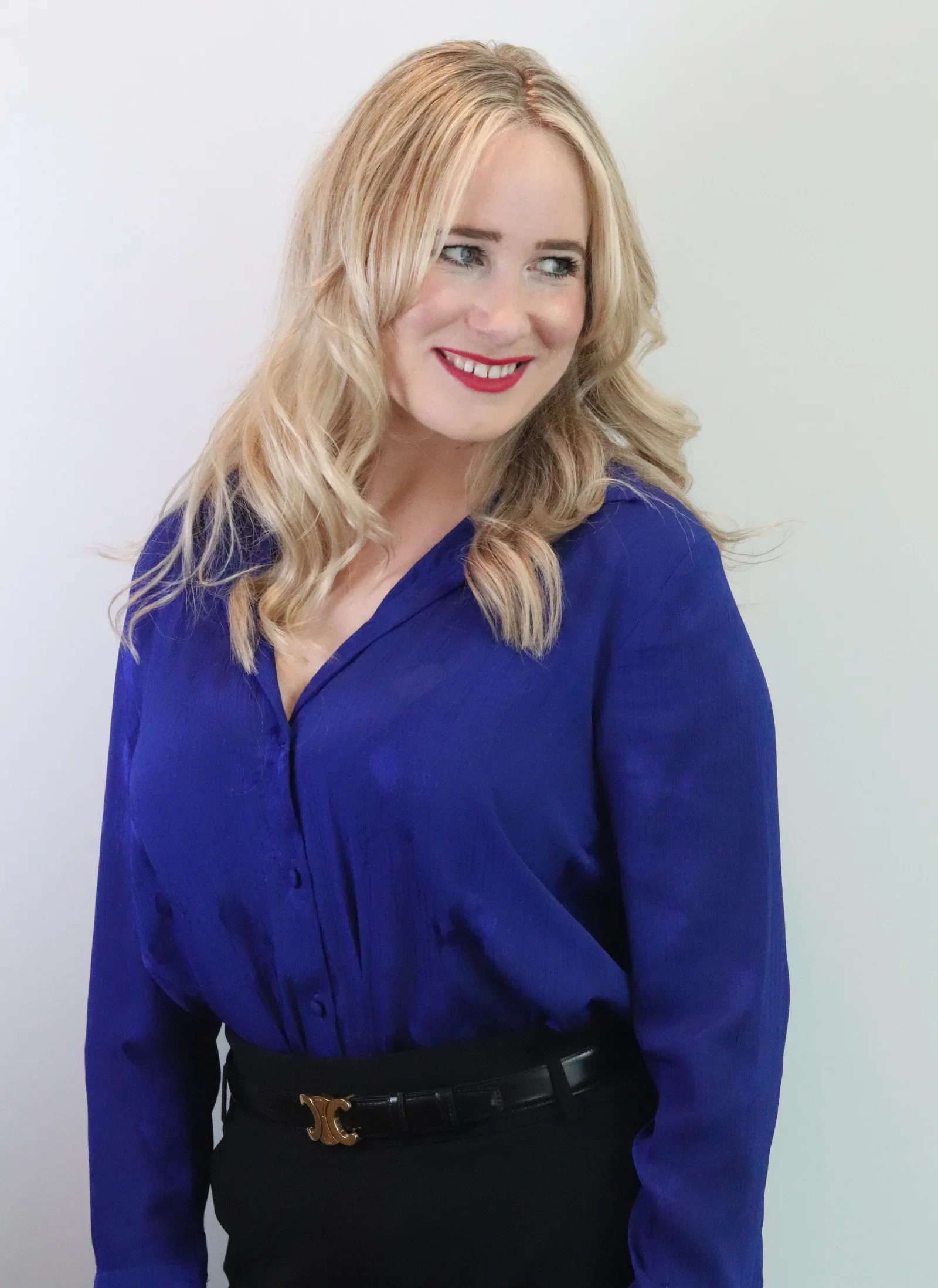 A woman with blonde hair, red lipstick, and a bright smile wearing a royal blue blouse and black skirt, standing against a plain white background.