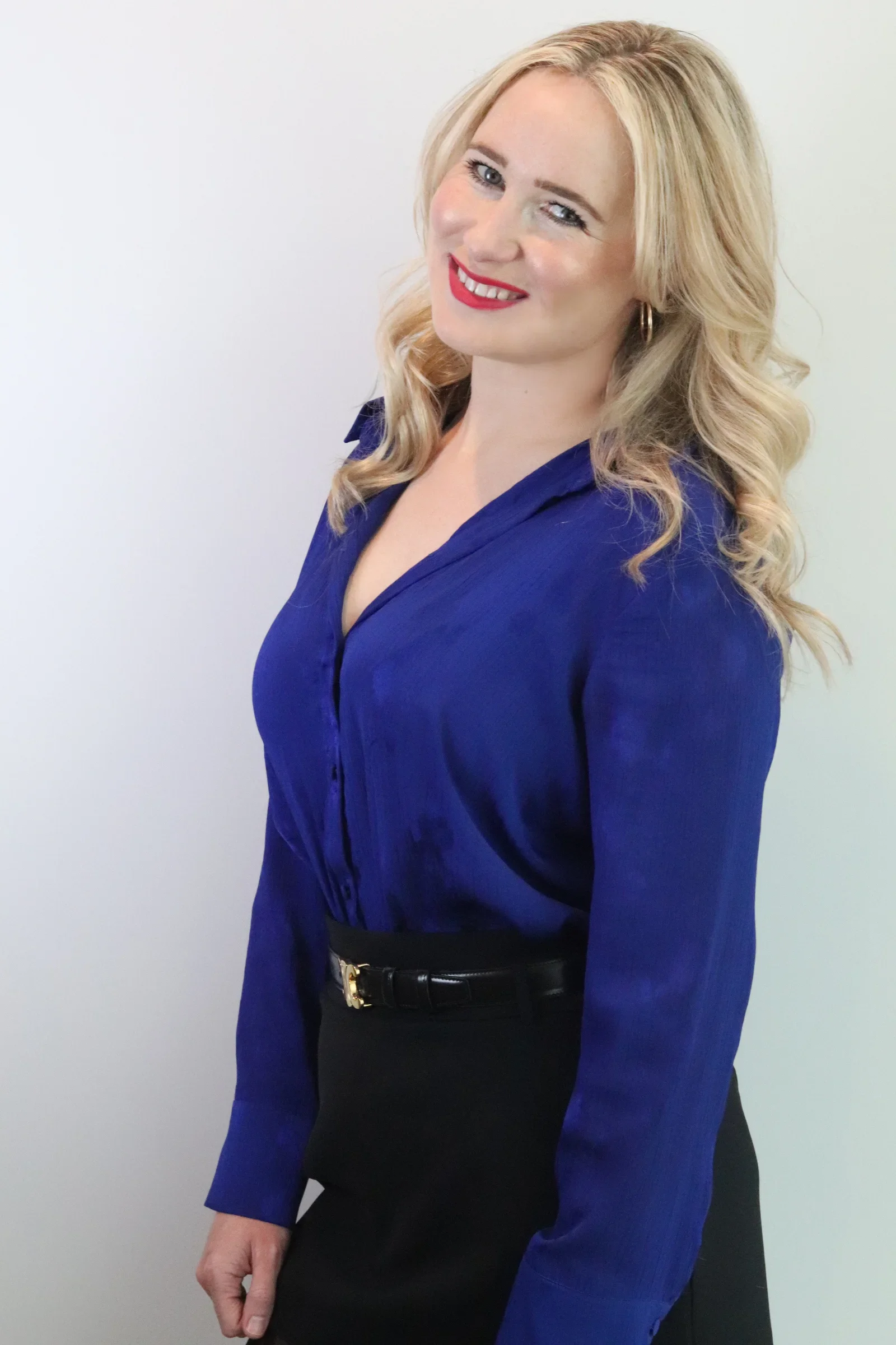 A woman with blonde, wavy hair, wearing a royal blue blouse and a black skirt, standing against a plain light-colored background and smiling.